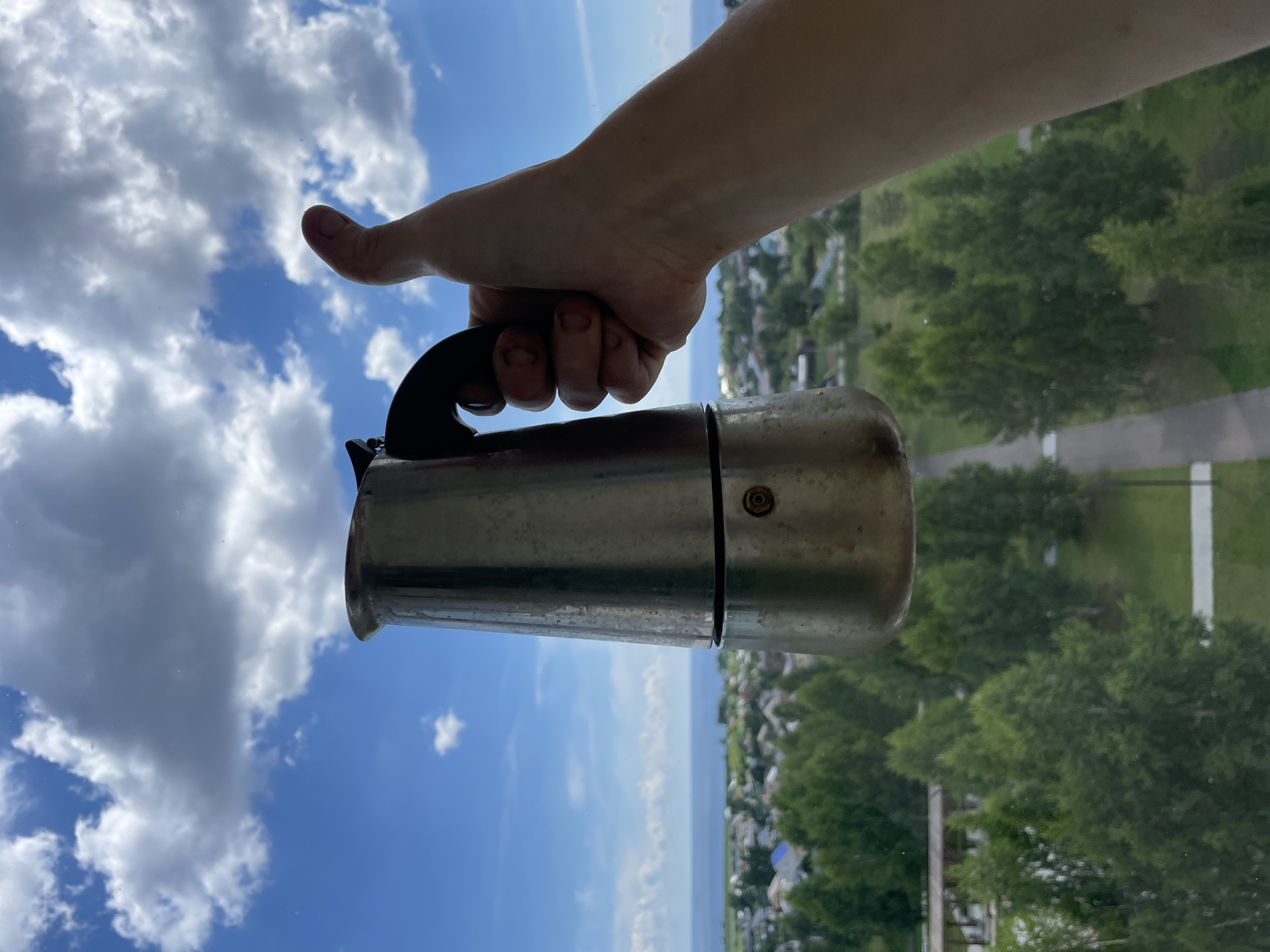 A person's hand holds a metal stovetop coffee maker in front of a window. The person's thumb is extended upwards in a "thumbs up" gesture. Outside the window, a blue sky with fluffy white clouds is visible. Below the sky, there is a body of water, likely the Kama River, followed by a low-lying area with green trees and houses, characteristic of Laishevo, Russia. A paved path can be seen winding through the greenery in the foreground. The coffee maker appears to be made of aluminum and shows some signs of use, with a black handle. The overall scene suggests a pleasant day with a view of a semi-urban landscape.