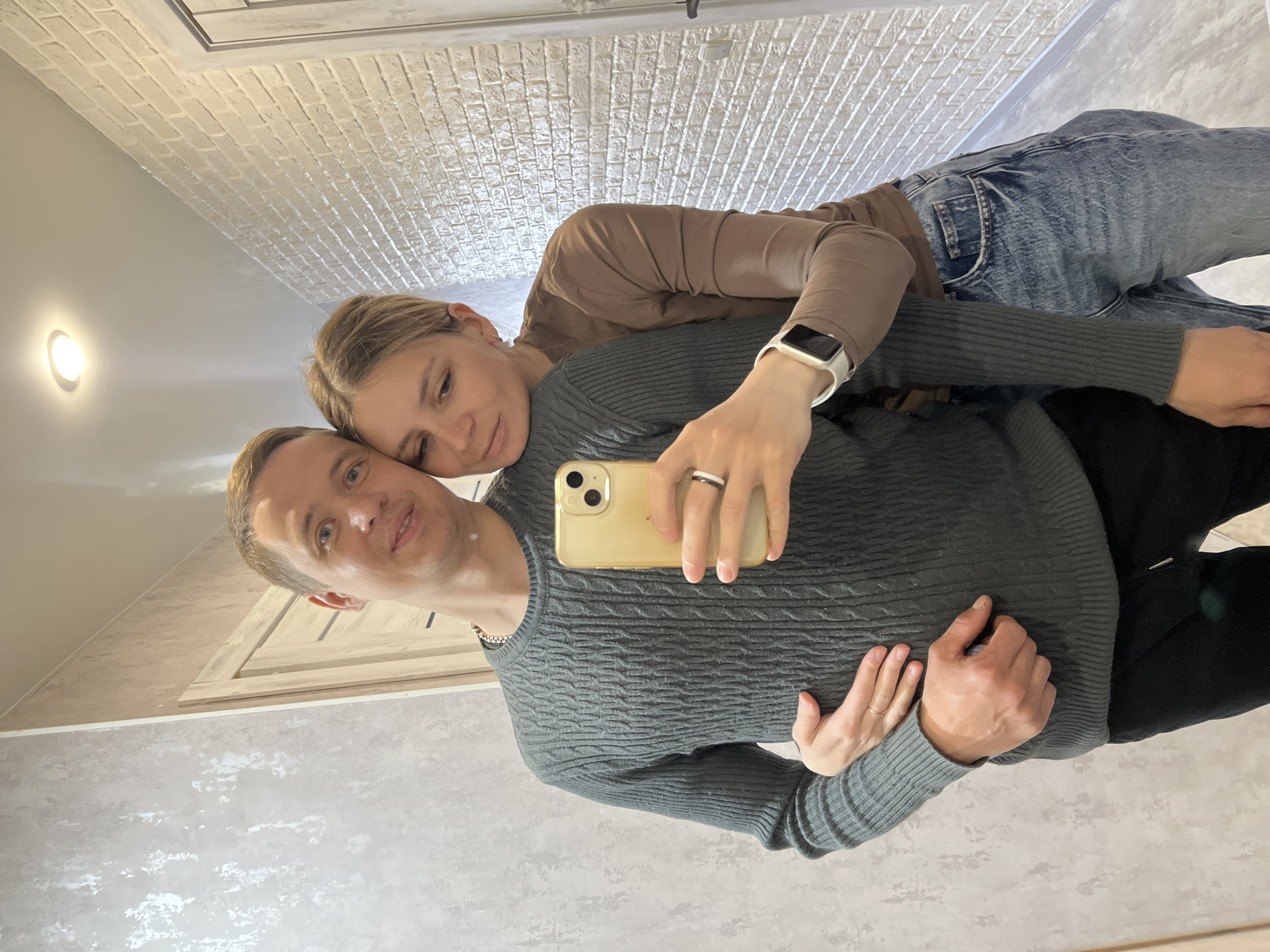 A selfie captures a man and a woman posing together in an indoor setting, likely a home in Chaikovsky, Russia. The image is taken in front of a mirror, with the man holding a gold-cased smartphone with three cameras. He is positioned in the foreground, facing the camera with a subtle smile. He has short, light brown hair and is wearing a dark grey, cable-knit sweater with a gold chain visible around his neck.
Behind him, the woman leans her head gently on his left shoulder, looking towards the camera with a soft, affectionate expression. She has blonde hair, possibly pulled back, and is dressed in a brown long-sleeved top and blue jeans. A white smartwatch is visible on her left wrist, and she wears rings on her right hand which is also holding the phone.
The background shows a brightly lit interior. To the right of the couple, a wall features a light-colored, textured brick pattern. To their left, another wall has a lighter, mottled grey or white finish. An oval-shaped light fixture is visible on this wall, emitting a warm glow. A light-colored wooden door frame is also discernible in the background. The overall mood is warm and intimate, conveying a sense of affection between the two individuals. There is no visible text in the image.