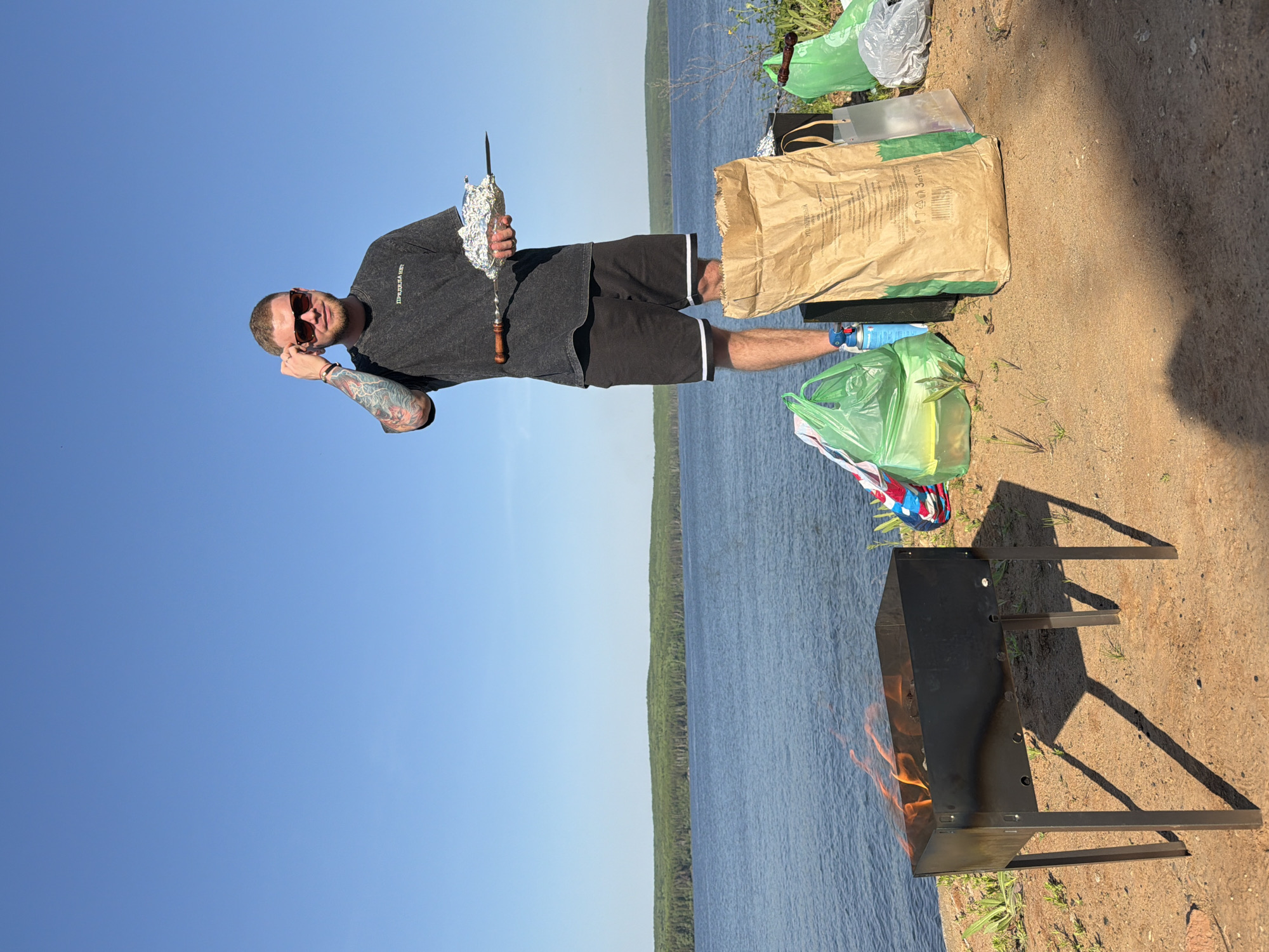 A man is standing next to a barbecue on the shore of a body of water. He is holding a skewer with food wrapped in foil. The barbecue has flames. There is a bag of something, and some trash bags nearby. A body of water and a treeline are in the background. The sky is visible. The picture was taken in the city of Chaikovsky, Russia.