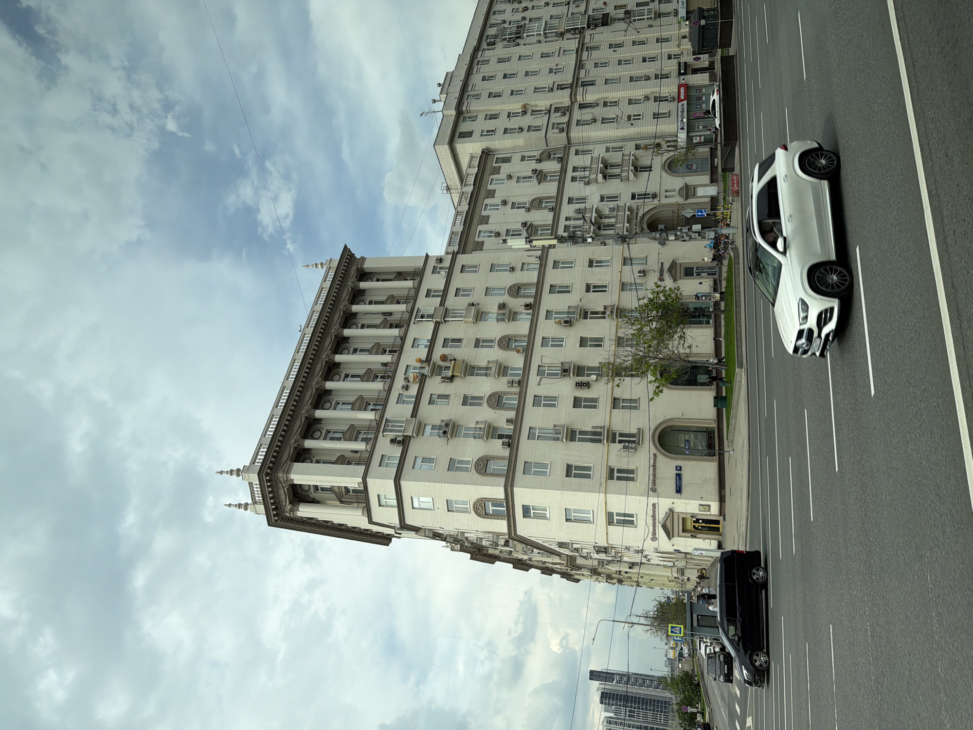 This image shows a view of a street in Moscow, Russia. In the background, there are large, multi-story buildings with a light-colored facade. There are cars driving on the multi-lane street. The sky is cloudy.