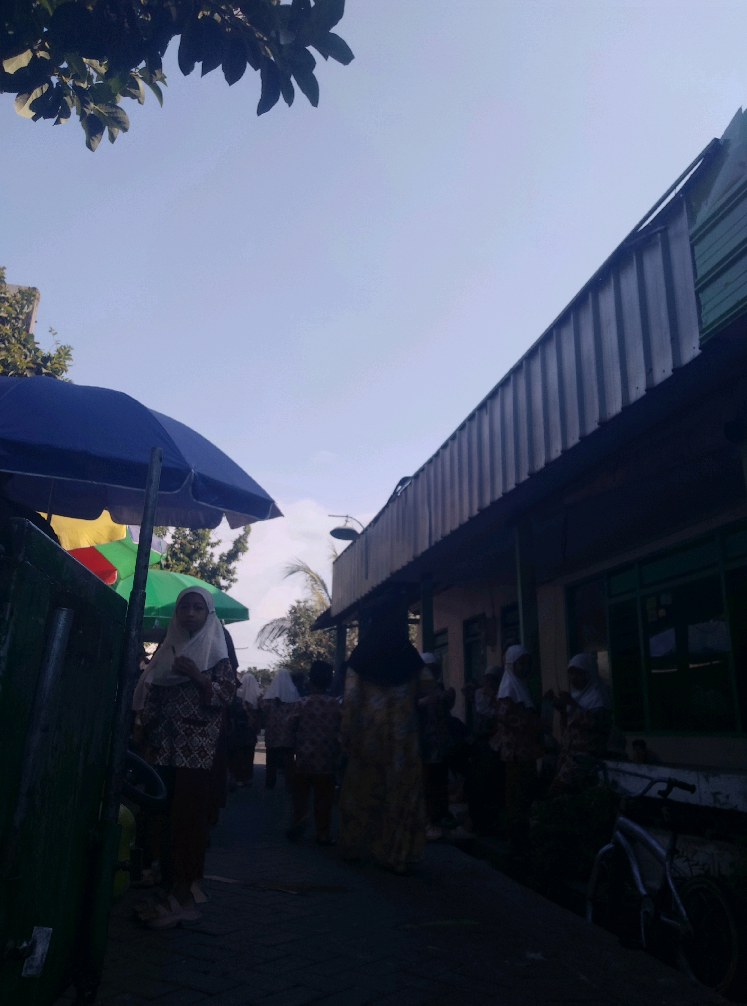 This image captures a street scene in Wotan, Indonesia. In the foreground, a young girl wearing a headscarf and a patterned dress stands by a dark green structure. Behind her, several other individuals, also in headscarves and patterned clothing, walk down a paved path. Colorful umbrellas, one blue and others partially visible in yellow and green, are set up to the left of the path. The background features a building with a corrugated metal roof and windows. Trees and a clear blue sky are visible above. A bicycle is parked near the building on the right. The lighting suggests it is daytime.
