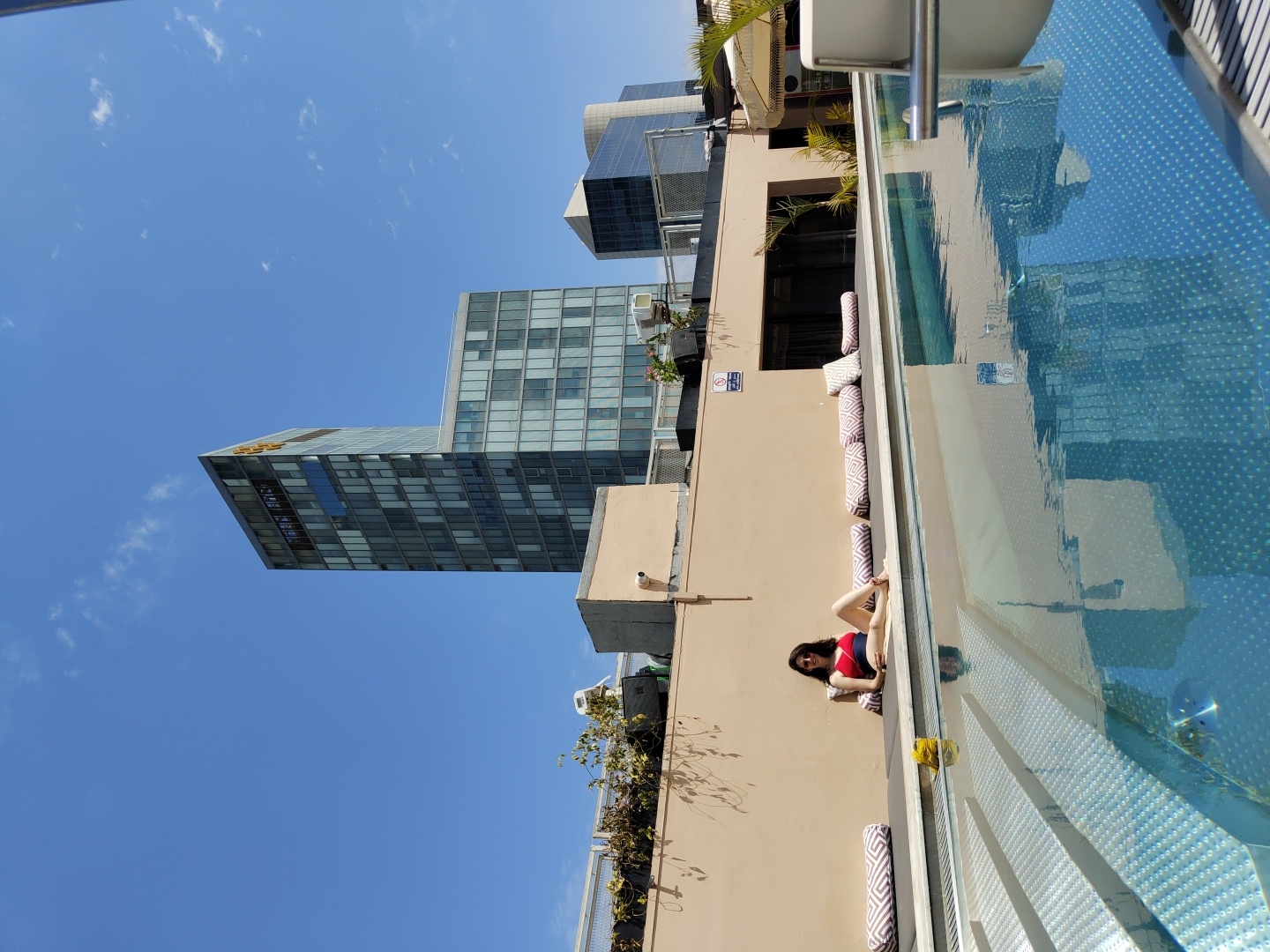 This vibrant daytime image captures a rooftop pool deck scene in Tel Aviv, Israel. The main subject is a woman in a red and dark blue swimsuit, lounging casually on a light brown wall adjacent to a swimming pool. She is positioned on her side, with her back mostly to the camera, appearing relaxed under the bright sun.
The sparkling blue swimming pool occupies a significant portion of the right side of the frame, reflecting the clear sky and nearby buildings. Several stacked lounge cushions, featuring a white and patterned grey design, are visible along the pool's edge, suggesting a comfortable seating area.
In the background, modern glass-clad high-rise buildings pierce a brilliant blue sky dotted with scattered white clouds. One prominent skyscraper features a distinctive golden text logo at its top, which appears to read "DAVID". Another dark-glass building is visible further back. The clear sky and strong sunlight indicate a sunny day, likely around midday.
Visible text includes a white circular sign on the beige wall, depicting a red circle with a crossed-out diving figure, clearly indicating "NO DIVING". Greenery is interspersed around the edges of the rooftop area. The scene conveys a sense of urban leisure and relaxation by a pool overlooking a modern cityscape.
