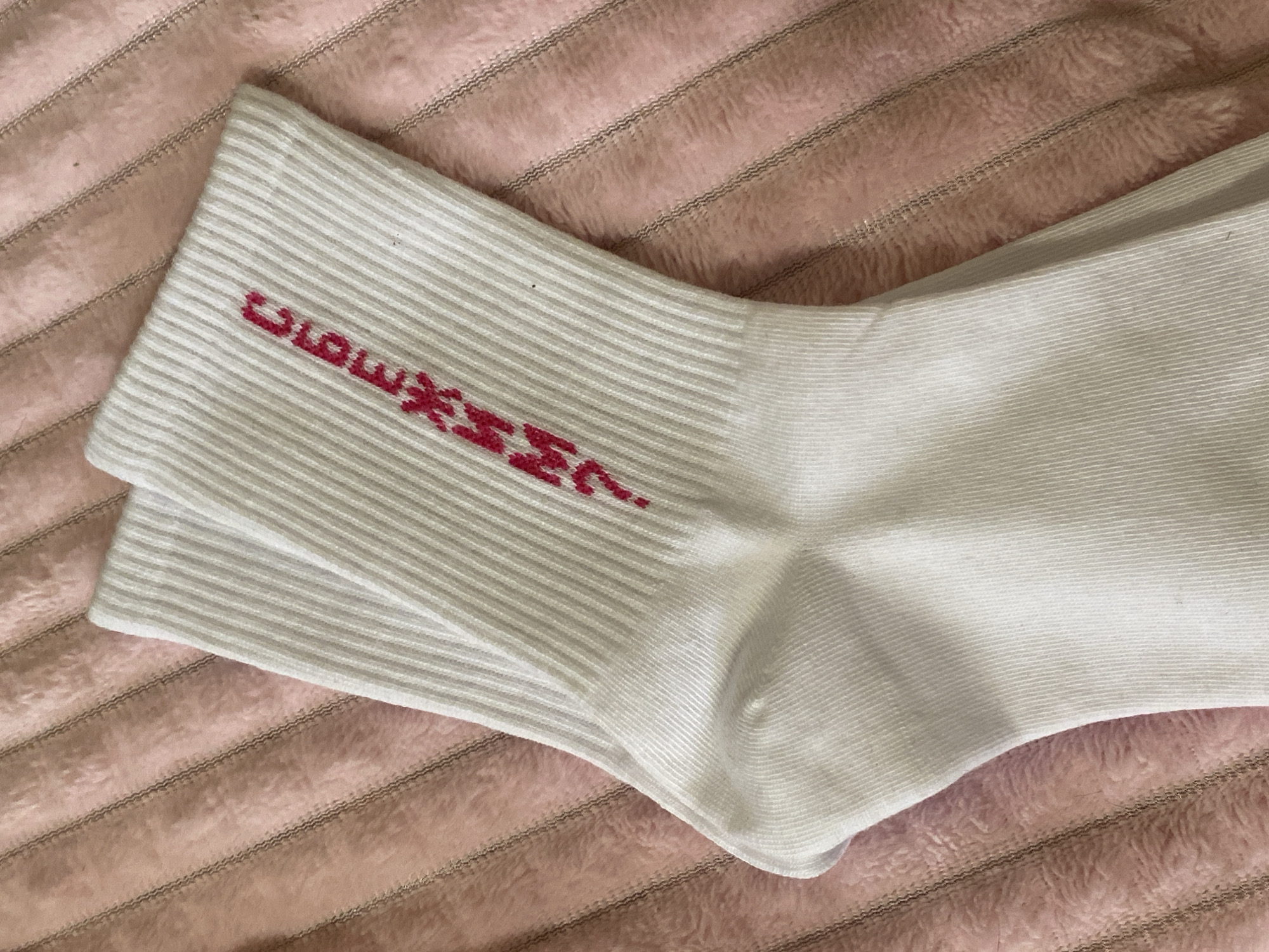 The image shows a pair of folded, white socks with thin, horizontal stripes. A red inscription is visible on the upper part of the socks. The socks are placed on a soft, pink, textured surface. There is no information to suggest a location in the city of Votkinsk, Russia, or anything related to it in this image.