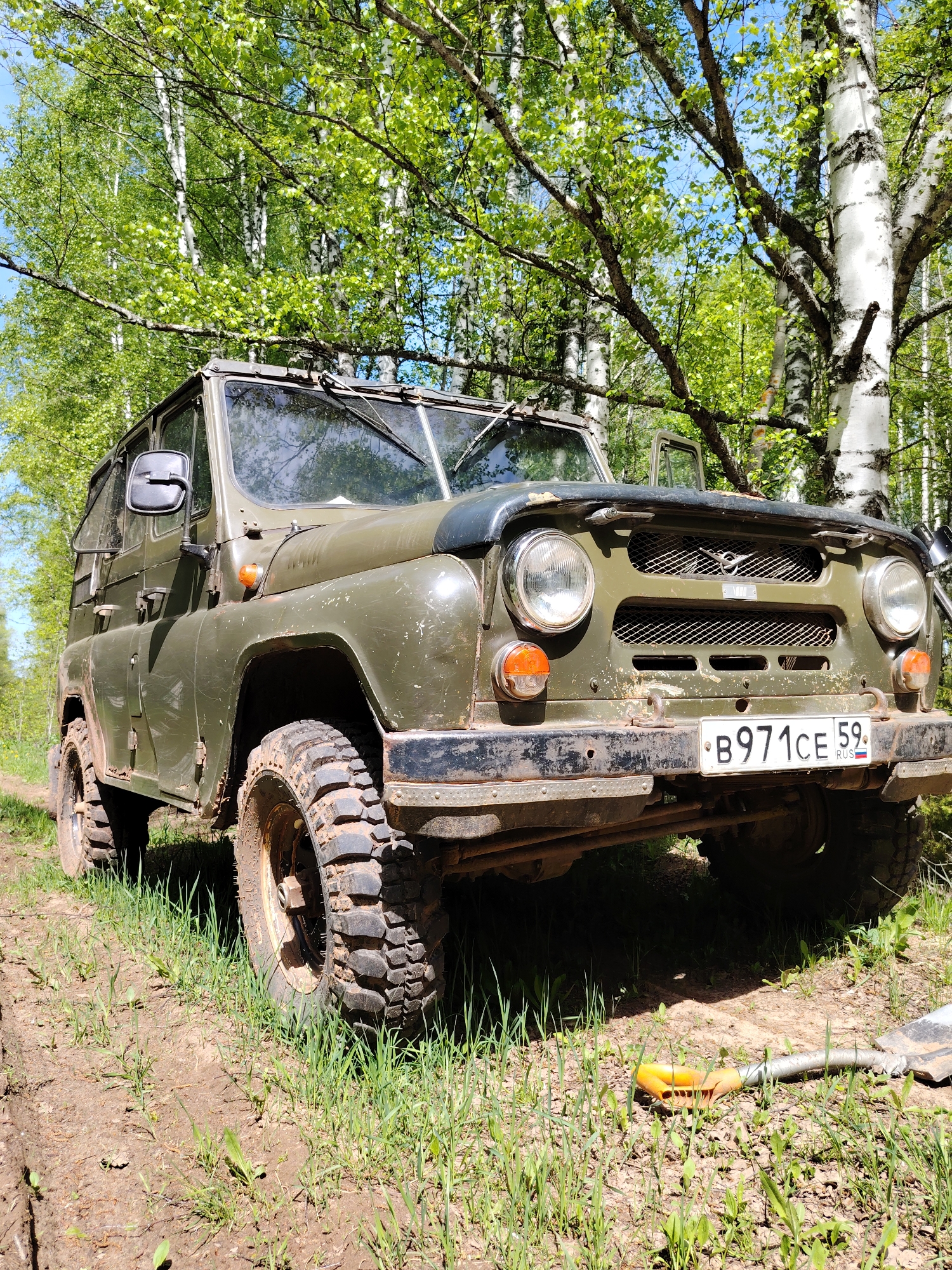 The image shows a green UAZ-469 military vehicle parked on a dirt road in a wooded area. The vehicle is facing forward and appears to be dirty. The front license plate reads "B971CE 59 RUS." A shovel lies on the ground to the right of the vehicle. Birch trees are visible in the background. The license plate indicates that the vehicle is registered in the Perm Krai region of Russia. There are no people visible in the image. The location appears to be in a forest in the city of Markovskii, Russia.