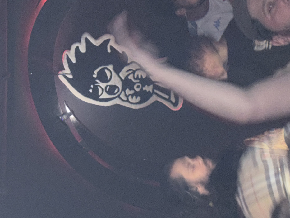 The image depicts an indoor, low-light scene consistent with a music venue or club. A prominent, stylized cartoon character logo, rendered in black with a white outline, is suspended against a dark, curved wall or ceiling. This character features spiky hair, an open mouth, wide eyes, and the letters "FTO" inscribed on its chest, holding a stick-like object. Red ambient lighting is visible along the curved architectural feature behind the logo. In the foreground and mid-ground, multiple individuals are present. On the left, a man with a beard and dark hair, wearing a plaid shirt, is partially visible and looking to the right. A raised arm, blurred by motion, occupies the central foreground. Other indistinct figures are visible in the background and to the right, including one wearing a dark t-shirt with short hair and another wearing a white t-shirt with a "Kappa" logo, indicating a gathering of people. The location is Le Trabendo, a music venue in Paris, France, identifiable by its distinctive FTO mascot.