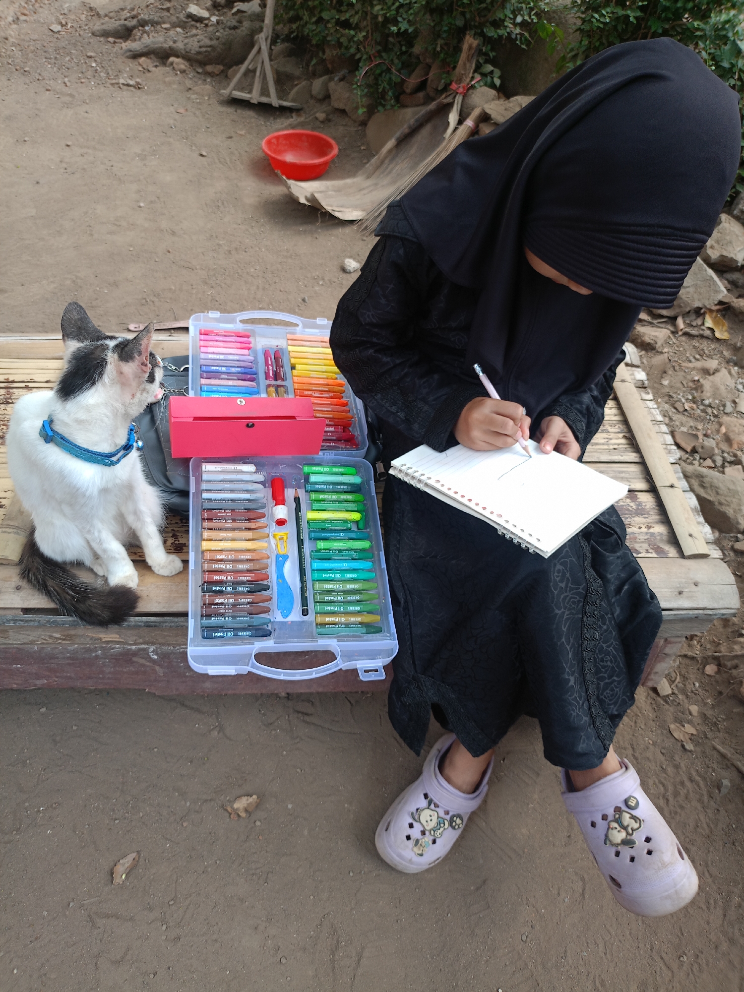 In Jelbuk, Indonesia, a young person wearing a black hijab and abaya sits on a wooden bench outdoors, drawing in a notebook with a pink pencil. Beside them, a black and white cat with a blue collar sits attentively. A large, clear plastic case filled with colorful oil pastels and art supplies is open on the bench. The ground is dirt, with scattered leaves and rocks. In the background, a red bowl and a broom leaning against a wall are visible.