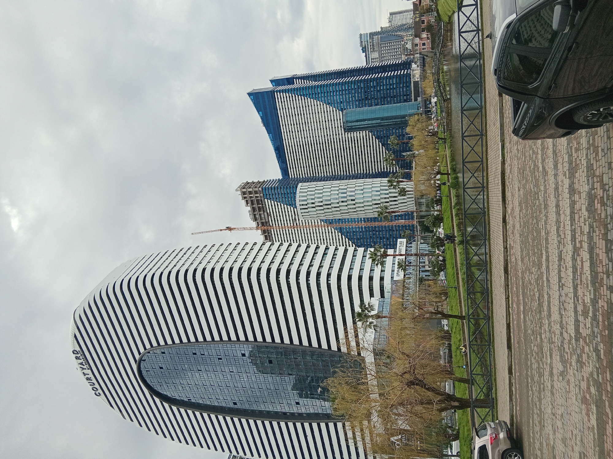 The image depicts a cityscape in Batumi, Georgia, under a cloudy sky. Prominently featured is the architectural landmark of the Courtyard by Marriott hotel, characterized by its distinctive white and black striped design with a large oval window. Adjacent to the hotel are several modern high-rise buildings, some partially clad in blue, indicative of ongoing construction. A construction crane is visible amidst these buildings. A water canal bordered by a black metal fence runs along the street, with sparse greenery and bare trees lining its banks. A paved walkway with brickwork pattern parallels the canal. A parked car is partially visible on the right side of the image, and another car is parked further down the street.