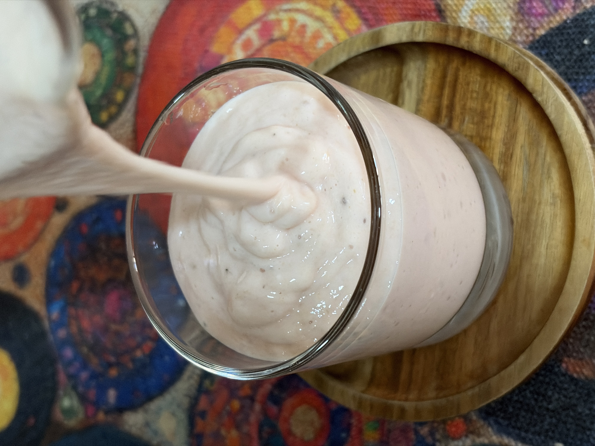 A glass of pink smoothie is being poured from another container into it. The glass is sitting on a round wooden tray. The background has a colorful circular pattern, and a woven texture. The specific location in Igboefon, Nigeria, cannot be determined from the image alone.