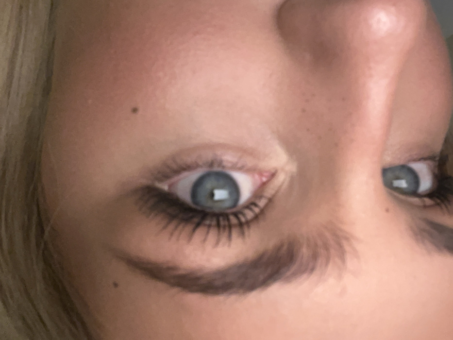 An extreme close-up of a face focuses on the eyes, nose, and eyebrows. The person has light skin, blue eyes, and dark brown eyebrows. They appear to be wearing mascara, enhancing the eyelashes. A mole is visible on the cheek. The camera angle is slightly tilted. The image has a soft, natural lighting.