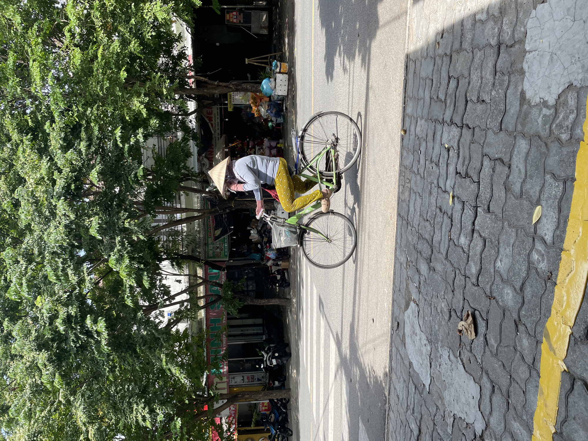 A person wearing a conical hat and a light blue long-sleeved shirt with pink wrist warmers, yellow pants with a white polka dot pattern, and tan sandals is riding a green bicycle with a basket on the front. The bicycle has a silver rack over the rear wheel and a blue seat. The person appears to be speaking on a cell phone. The bicycle is on a paved road with a pedestrian crossing marked by white stripes in the background. To the left of the road, there are shops with motorbikes parked in front and trees providing shade. The foreground consists of a grey cobblestone sidewalk with a yellow painted line. The scene is set in Đà Nẵng, Vietnam, under a bright, sunny sky, casting shadows on the road and sidewalk.
