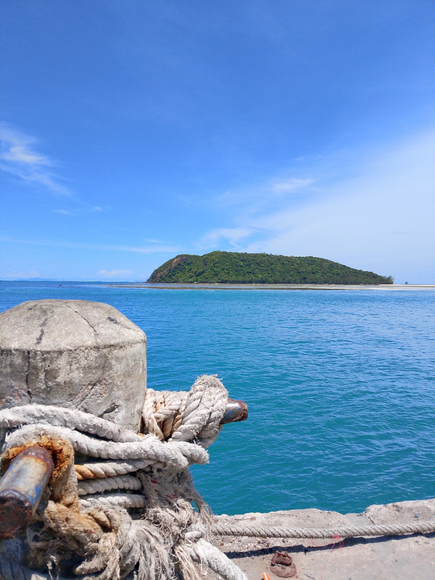 The image depicts a bright, sunny day with a clear blue sky over a tranquil turquoise sea. In the foreground, a weathered concrete bollard is prominently featured, secured with thick, worn ropes. The ropes are frayed and discolored, suggesting exposure to the elements and heavy use. A rusty metal ring is attached to the bollard, also wrapped with rope. The foreground also shows a concrete pier. In the background, a lush green island with a rounded peak rises from the water, its slopes covered in dense vegetation. The island is bordered by a sandy shoreline and shallow waters. The overall scene evokes a sense of a peaceful tropical harbor or coastline, likely in the vicinity of ตำบลเกาะพะงัน, Thailand, given the characteristic island landscape and clear, warm waters. There are no people visible in the image.