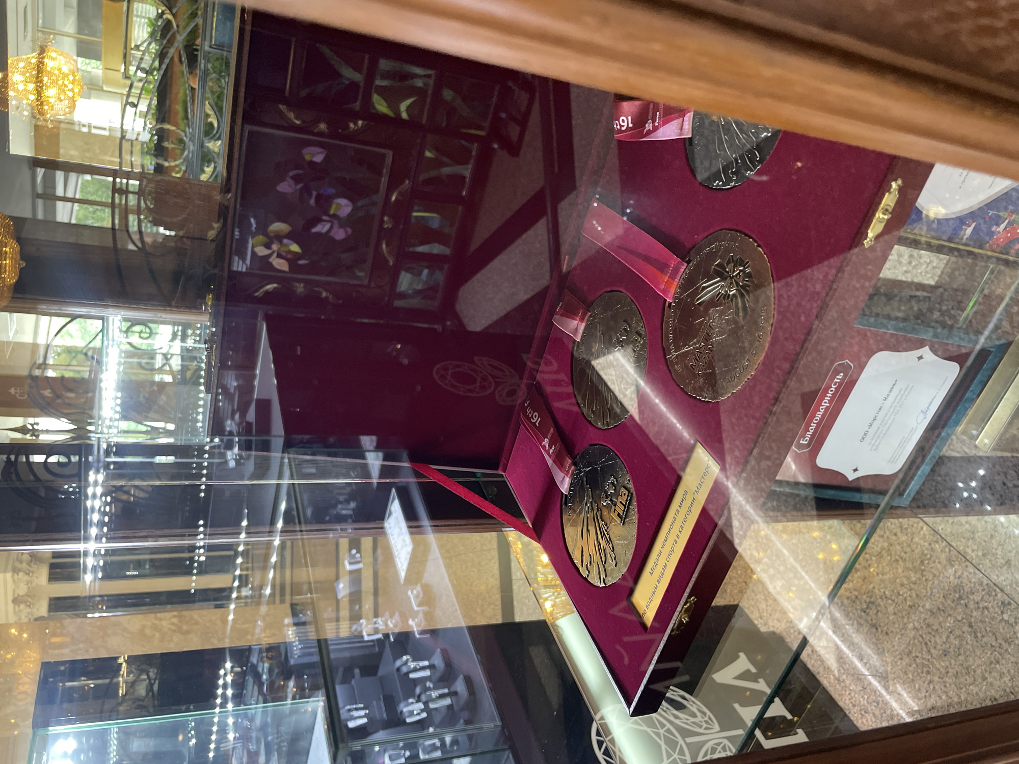 The image shows a display case with medals and a certificate. The medals are gold and silver, and one is engraved with "FINA MASTERS KAZAN AUGUST 5-16, 2015". The certificate is in Russian and reads "Благодарность" (Gratitude). The display case appears to be in a public building, possibly a museum or exhibition hall, given the presence of other display cases in the background. The lighting suggests an indoor setting. The certificate's mention of Kazan, Russia, indicates the location.