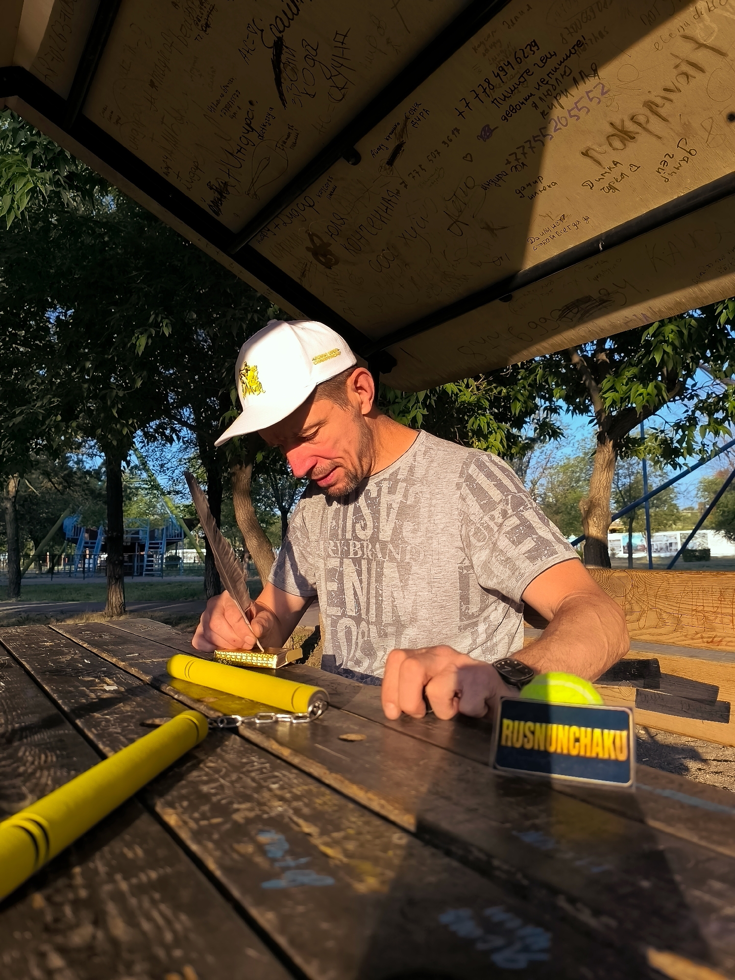 An outdoor scene shows a man sitting at a wooden picnic table. He wears a white baseball cap with a yellow emblem and a grey t-shirt. His gaze is directed downwards at the table where he holds a quill pen in his right hand, appearing to write on a small object. In front of him on the table are yellow nunchaku. To his right is a bright yellow ball and a dark rectangular object with the word "RUSNUNCHAKU" in yellow lettering. The table is weathered and covered with faint markings. Behind the man, trees with lush green foliage are visible, and in the distance, playground equipment can be seen. The sky is a clear blue. The lighting suggests it is late afternoon, with sunlight casting long shadows. The overhead structure of the picnic area is covered in graffiti.