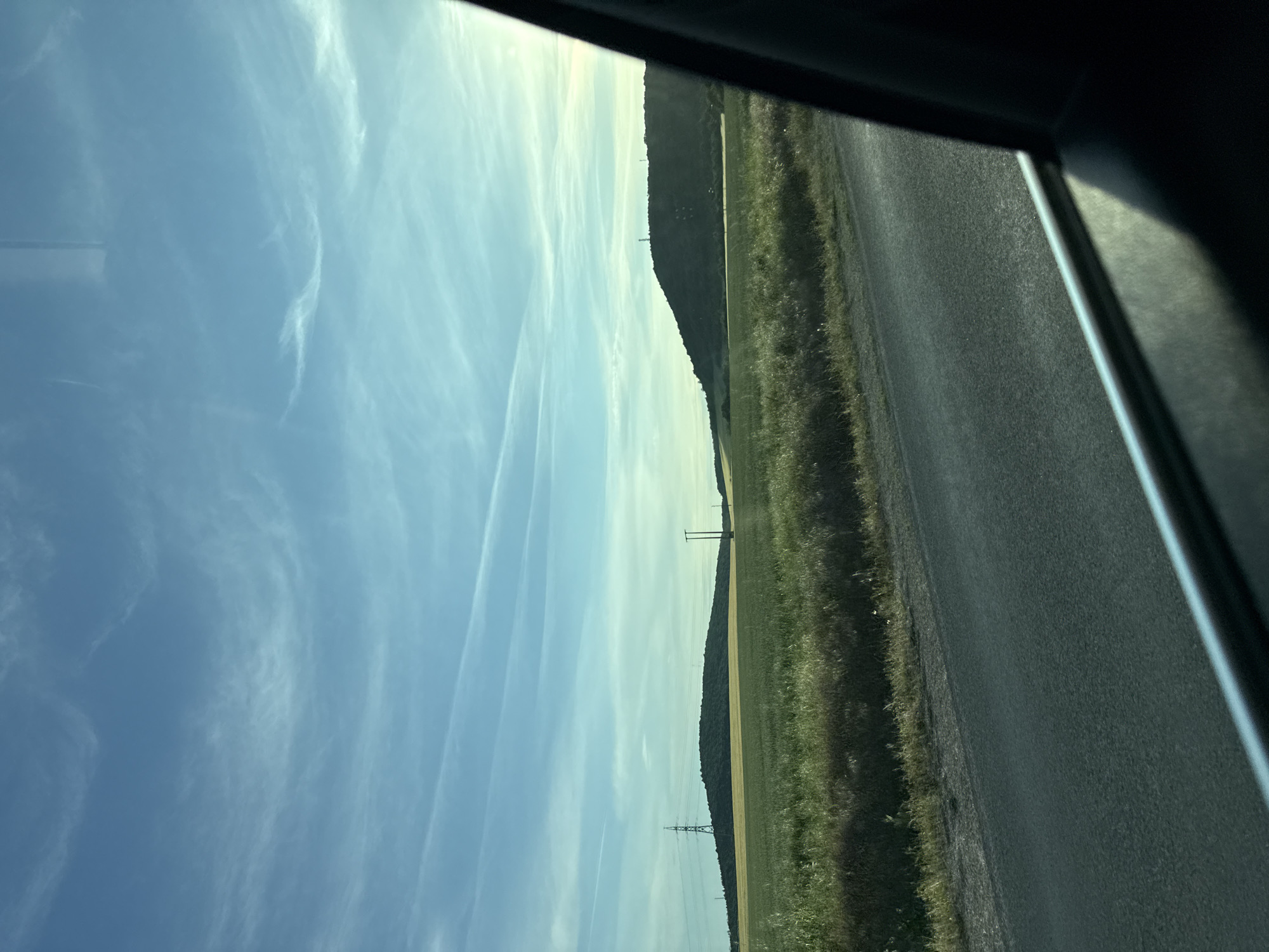 The image shows a view from inside a vehicle, likely a car, looking out the window. The scene is a rural landscape. A clear blue sky with wispy contrails fills the left side of the frame. A field and a line of trees run horizontally across the middle of the image. Electrical pylons are visible in the field. A paved road occupies the right side of the frame. The car's interior trim is visible at the edges of the image. There are no people visible. The location cannot be determined from this image, nor is it possible to confirm if the image was taken in Slavyani, Bulgaria.