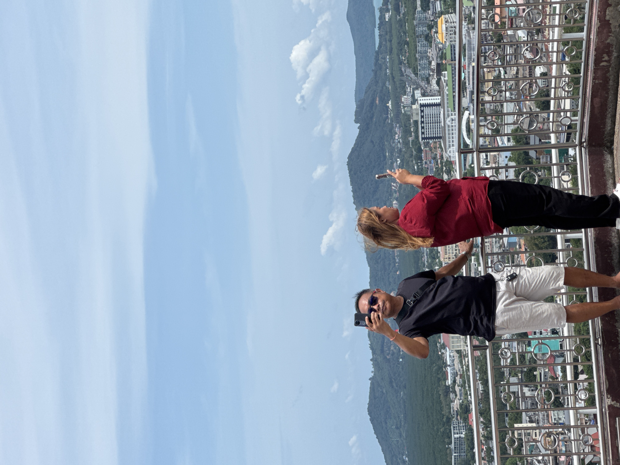 Two individuals are standing on an elevated platform, a man on the left and a woman on the right, both taking photographs with their mobile phones. The man is wearing a black t-shirt, light-colored shorts, and sunglasses, with a sling bag across his chest. The woman is dressed in a red long-sleeved top and black pants. They are positioned behind a metal railing overlooking a city with buildings and lush green hills in the background, under a partially cloudy blue sky. The scene appears to be a scenic viewpoint in Ratsada, Thailand, likely offering panoramic views of the city and its surroundings.