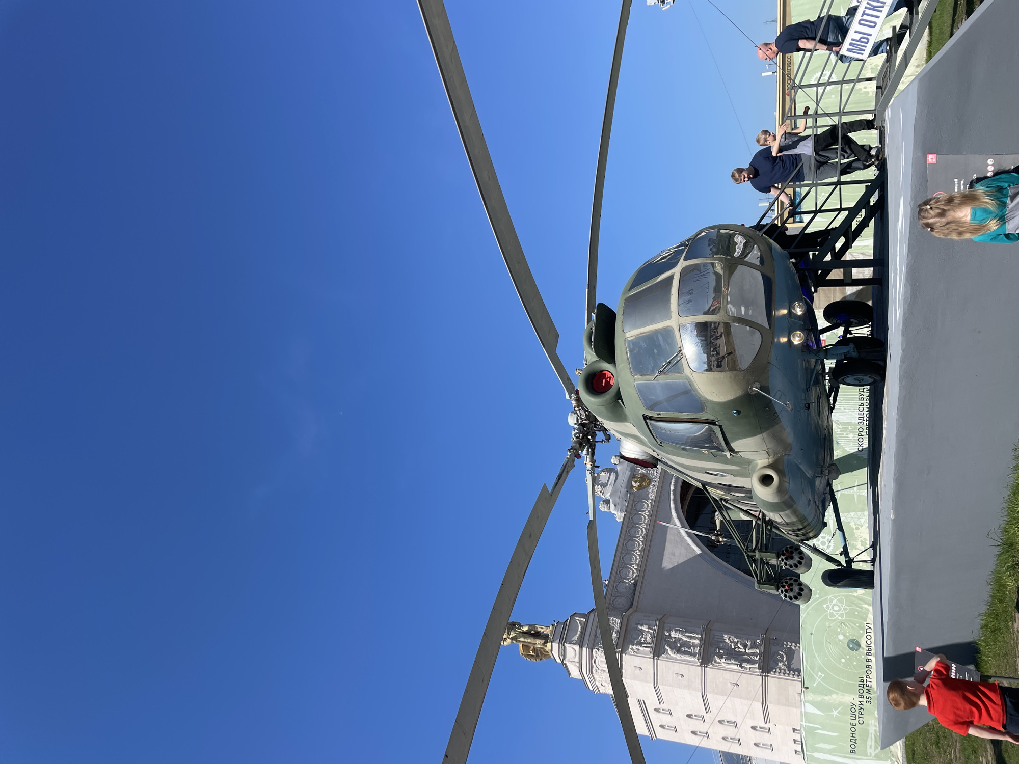 The image shows a Mil Mi-8 helicopter on display in Moscow, Russia, with visitors observing it. The helicopter, painted in camouflage green and grey, is positioned on a grey platform near a building with ornate architectural details, including sculptures. The helicopter's rotor blades extend towards a clear, blue sky.
Several people are visible: a group of adults and children are looking at the helicopter from a raised platform or staircase, and a child in a red shirt stands near a display board with text written in Russian. The display board features information about a water show with 35-meter high water jets, suggesting that the helicopter is located near an entertainment or exhibition area.
Overall, the scene depicts a typical outdoor exhibit in Moscow, with visitors engaging with the displayed helicopter and nearby attractions.