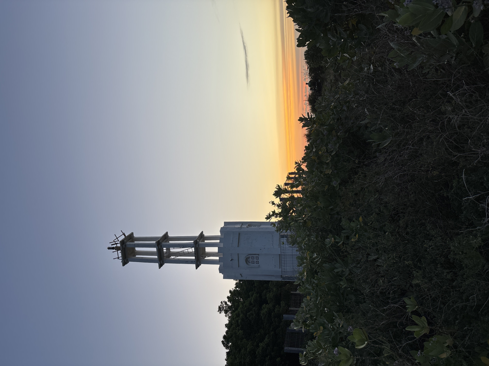 The image captures a low-angle view of a white tower structure, partially obscured by dense green vegetation, against a backdrop of a gradient sky that transitions from a light blue hue at the bottom to a golden-orange at the top, possibly indicating sunset or sunrise. The tower appears to be composed of a solid white section with window-like detailing, topped with an open framework of ladders and supports. The location is Pecatu, Indonesia.