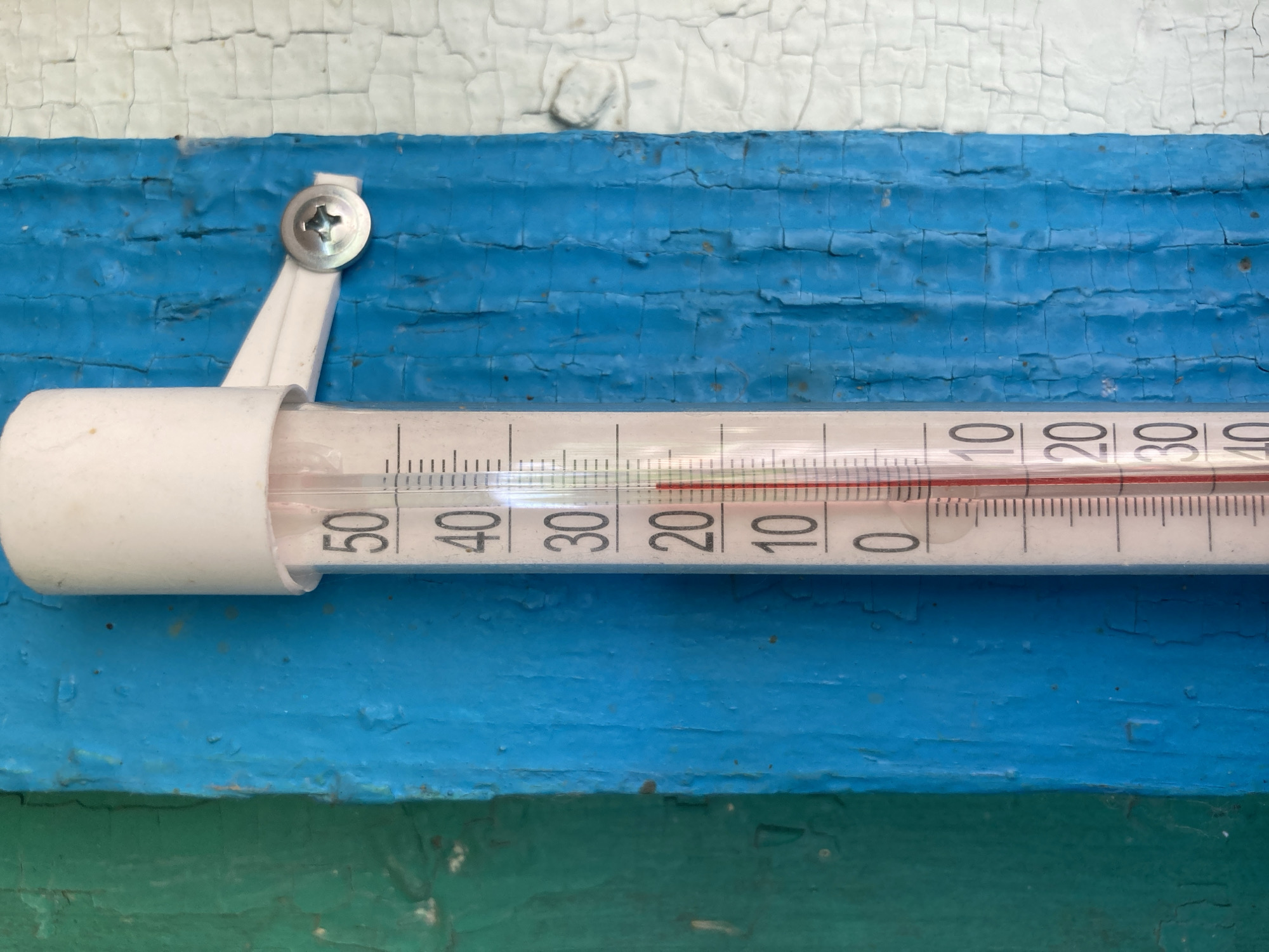 A close-up, eye-level shot shows a thermometer mounted on a weathered wooden surface painted in alternating vertical stripes of green, blue, and white. The thermometer itself is a glass tube with a white plastic cap and a white bracket secured by a silver screw. The glass tube displays a temperature scale ranging from 0 to 50 degrees Celsius, with markings for every degree and larger markings every ten degrees. A red liquid column within the thermometer indicates the current temperature, which appears to be between 20 and 30 degrees Celsius, closer to 27 degrees. The wooden surface exhibits cracks and peeling paint, suggesting outdoor exposure and age. The background is predominantly the thermometer and the painted wood. The scene is devoid of people or any visible action.