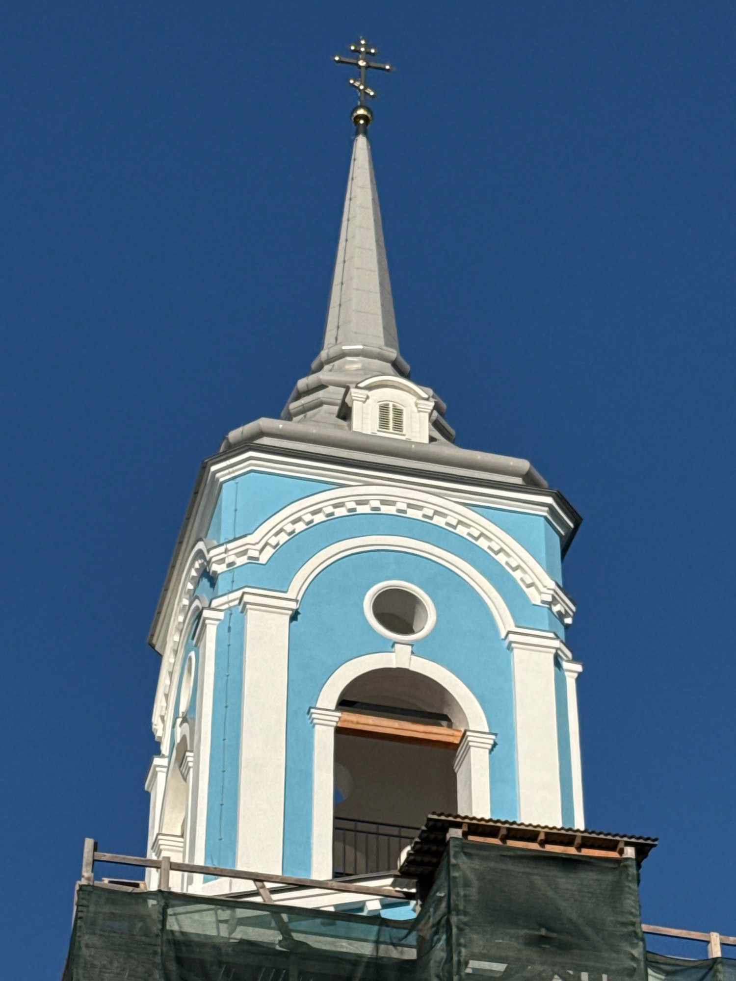 The image shows the top of a church tower against a clear blue sky. The tower is painted in bright blue and white with ornate detailing, featuring arched openings and decorative trim. At the very top, a slender, grey spire is topped with a golden Orthodox cross. The sky is a deep, uniform blue, suggesting it is daytime with good weather.
Part of the tower appears to be under construction or renovation. Scaffolding and dark green netting are visible at the base of the frame, covering some of the lower sections of the structure. Wooden beams and framing elements are also apparent, indicating ongoing work.
The architecture suggests a Russian Orthodox church, consistent with the provided location context of Valday, Russia. The bright colors and detailed ornamentation are characteristic of religious buildings in the region. The overall impression is one of a historic and significant religious edifice, possibly undergoing restoration to preserve its beauty. No people are visible in the frame, and no text is discernible. The time of day appears to be midday or early afternoon due to the clear, bright lighting.