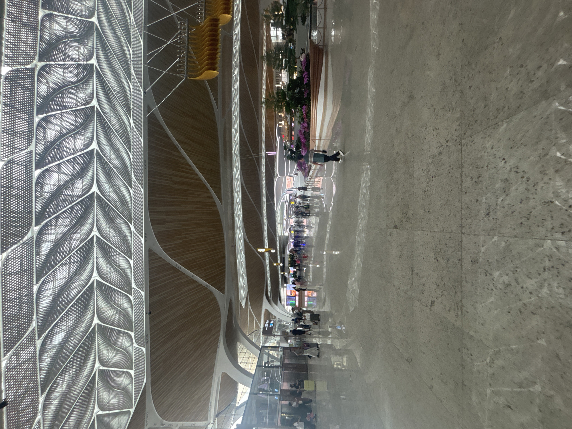 The image depicts a wide, polished marble floor of an airport terminal in Baiyun District, China. The ceiling features an intricate, wave-like design with metallic accents and wood paneling. Various people are walking through the terminal, some with luggage. In the mid-ground, a large, decorative planter with pink flowers and greenery is situated. Further in the distance, there are information displays and architectural elements typical of a modern airport. The lighting appears to be a mix of natural light filtering through the ceiling and artificial lights.