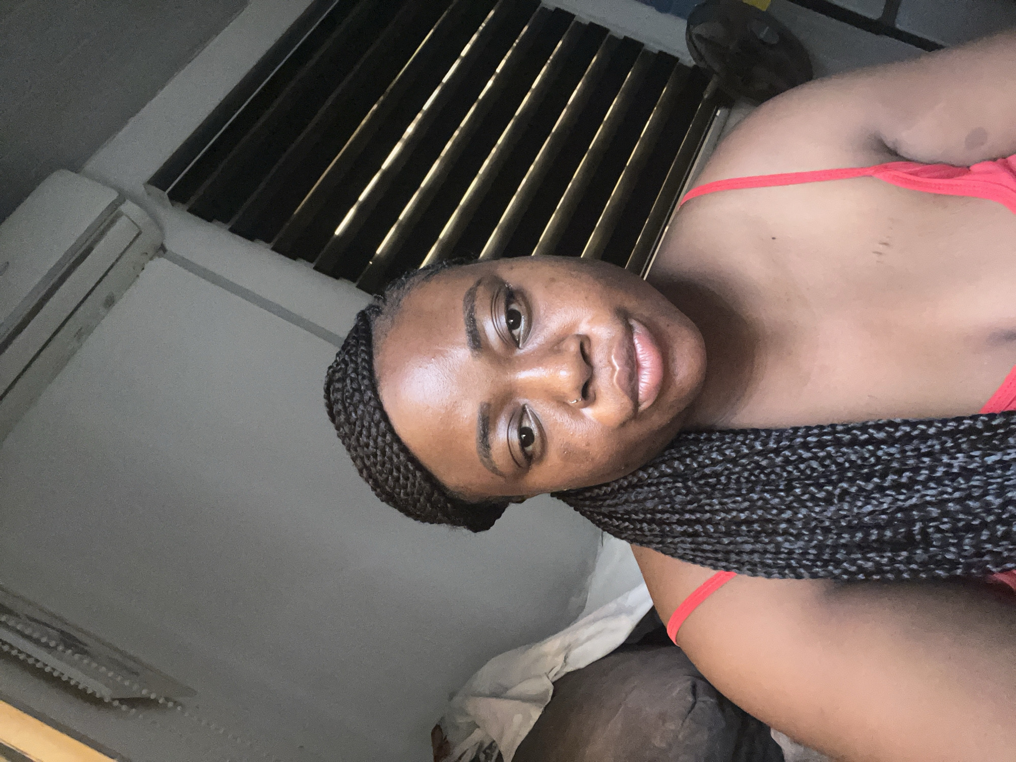 The image features a person with dark skin, wearing braids and a red top. The person is looking at the camera. The background includes a window with blinds and an air conditioning unit. The location is possibly in Abuja, Nigeria.
