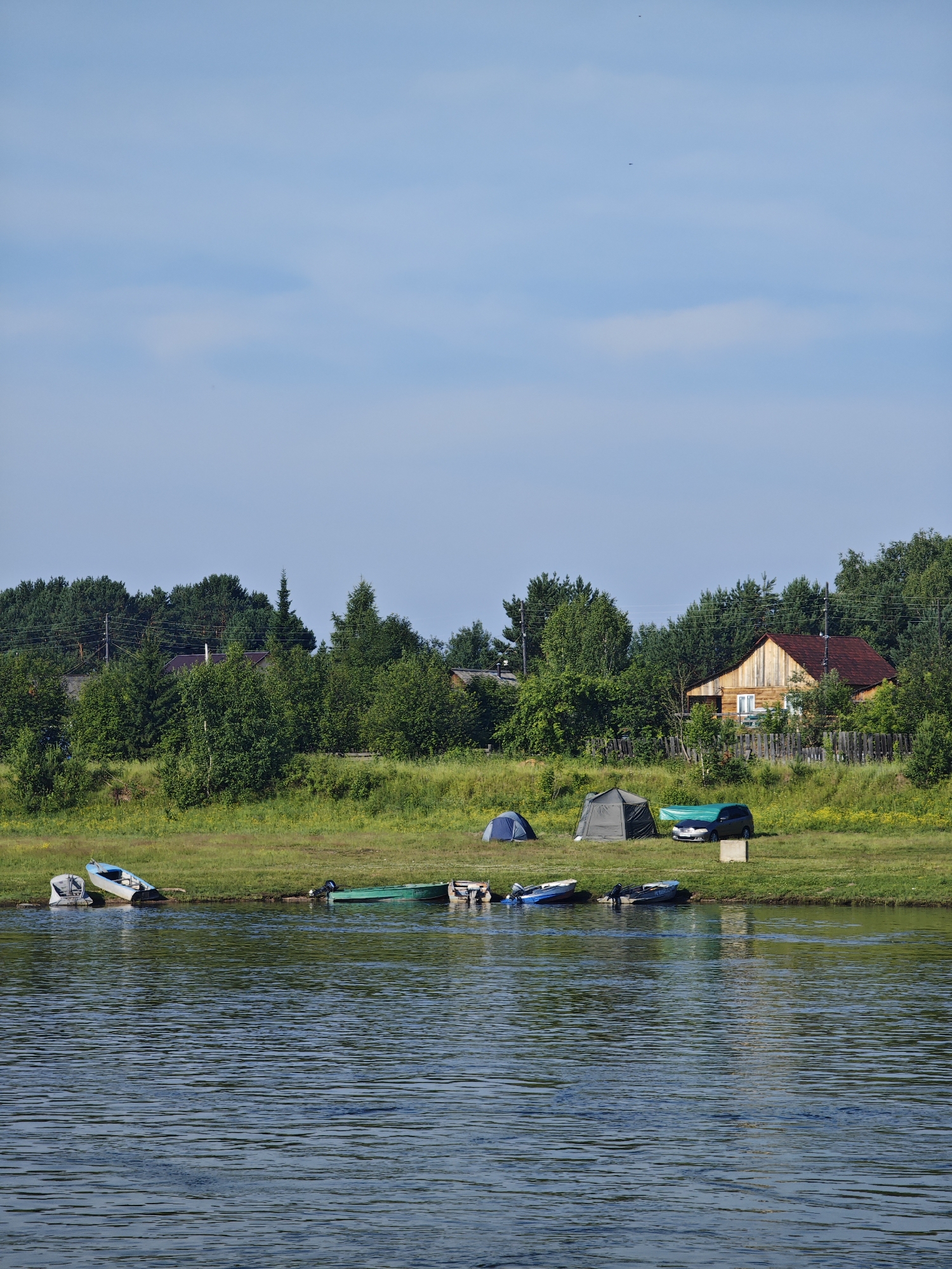 The image depicts a rural landscape, likely near the village of Galanino, Russia, characterized by a wide river in the foreground and a treeline with scattered houses in the background. Several small boats are moored along the grassy riverbank. On the shore, two tents and a dark-colored car covered with a green tarp are visible, suggesting a camping or recreational activity. The sky is a clear blue with a few wispy clouds. The overall scene is serene and suggests a peaceful, natural setting.