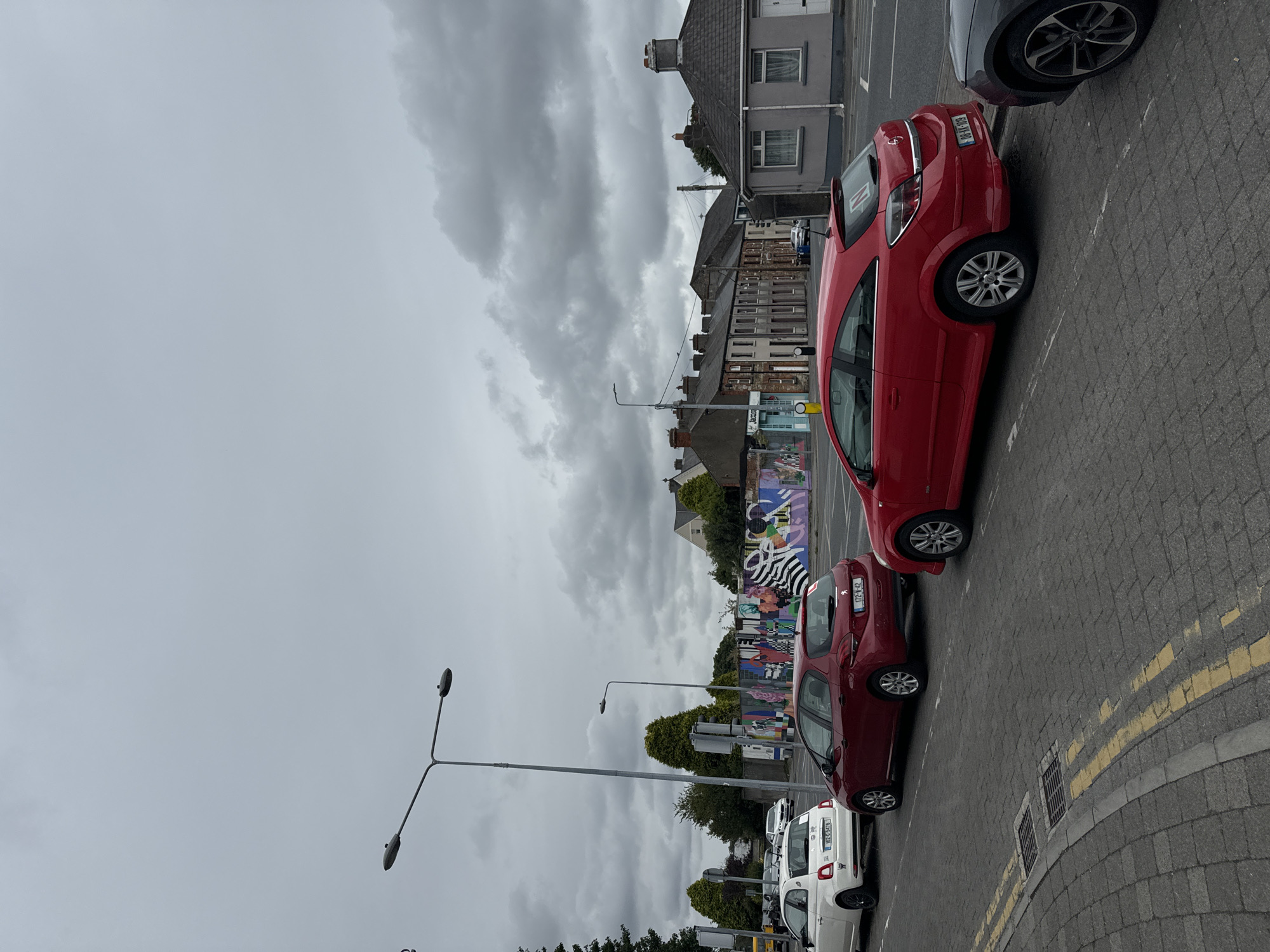 The image shows a street scene in Waterford, Ireland, under a cloudy sky. In the foreground, several cars are parked along the side of the road, including a white Fiat 500, a red Peugeot 208, and a red Opel Astra. The road surface is paved with cobblestones, and yellow lines mark the edge of the parking area. In the background, a mural covers a wall, depicting various colorful patterns and shapes. Residential buildings and streetlights line the street. The overall impression is a typical urban environment on an overcast day.