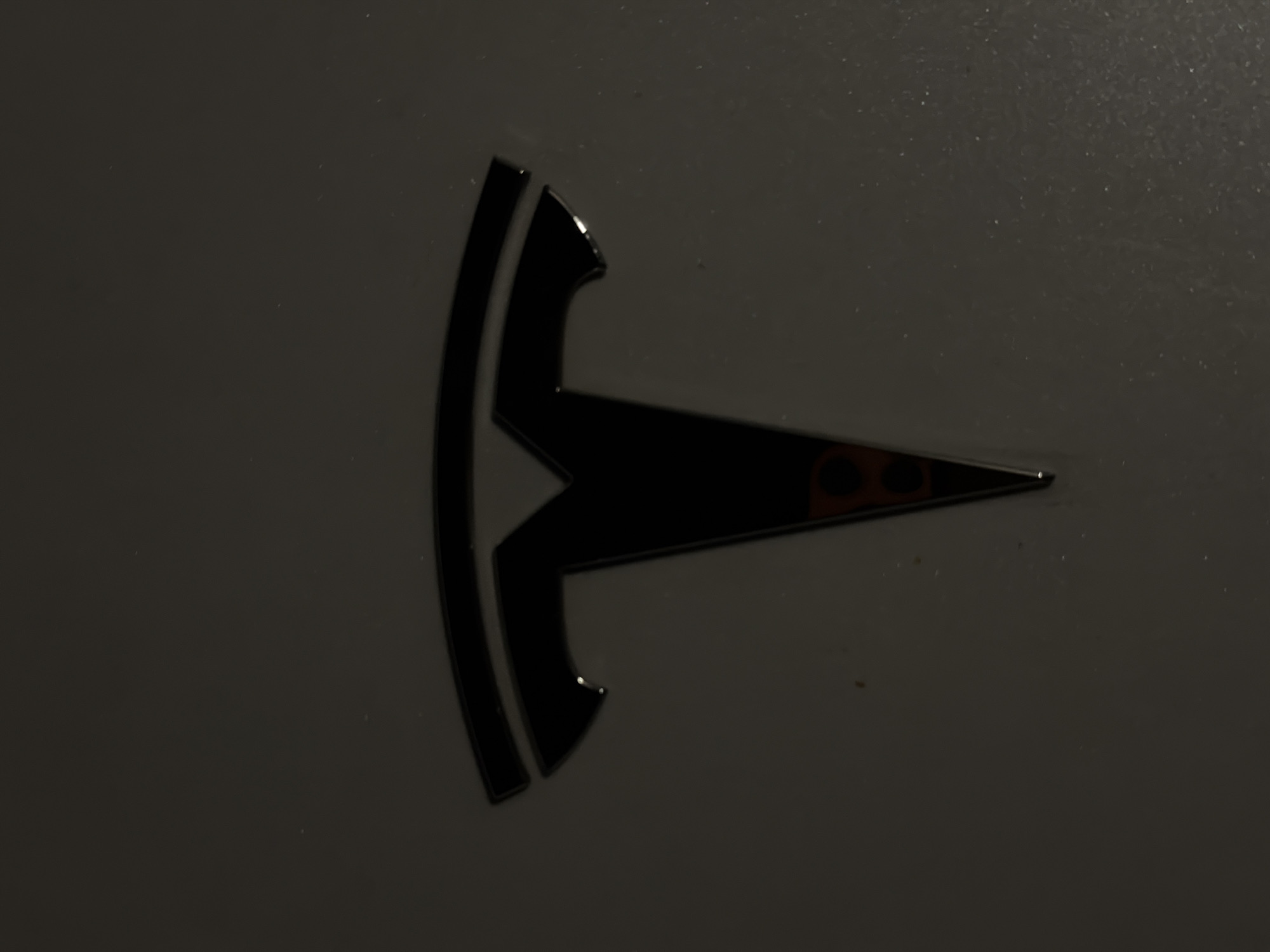 The image shows a close-up of a Tesla logo, which is a stylized "T" with curved lines above it. The logo is black and appears to be made of metal. It is mounted on a dark gray surface, possibly a car. There are two small red circles within the lower part of the "T". The lighting is dim, creating shadows and reflections on the surface of the logo and the background. The image does not provide any information about the location in the city of Bila Tserkva, Ukraine.