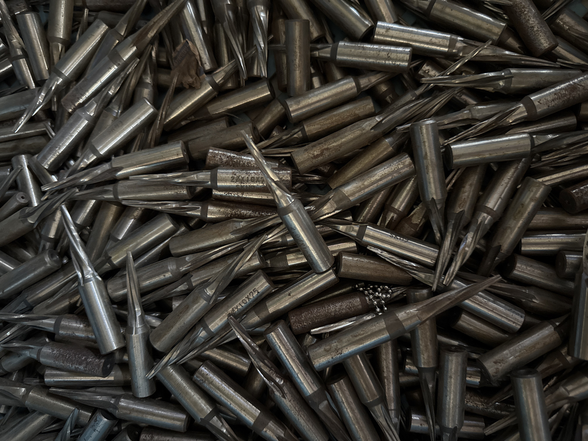 A close-up, overhead shot displays a large pile of used, metallic soldering iron tips. The tips are of various sizes and shapes, with some showing signs of wear and corrosion. They are densely packed together, with their pointed ends facing in multiple directions, creating a chaotic and textured surface. The lighting is dim, casting shadows and highlighting the metallic sheen of the objects. There are no people, animals, or any recognizable elements of a city environment, such as buildings or streets, visible in the image. The scene is devoid of any specific location markers, making it impossible to determine its presence in Chalong, Thailand or any other geographical location.