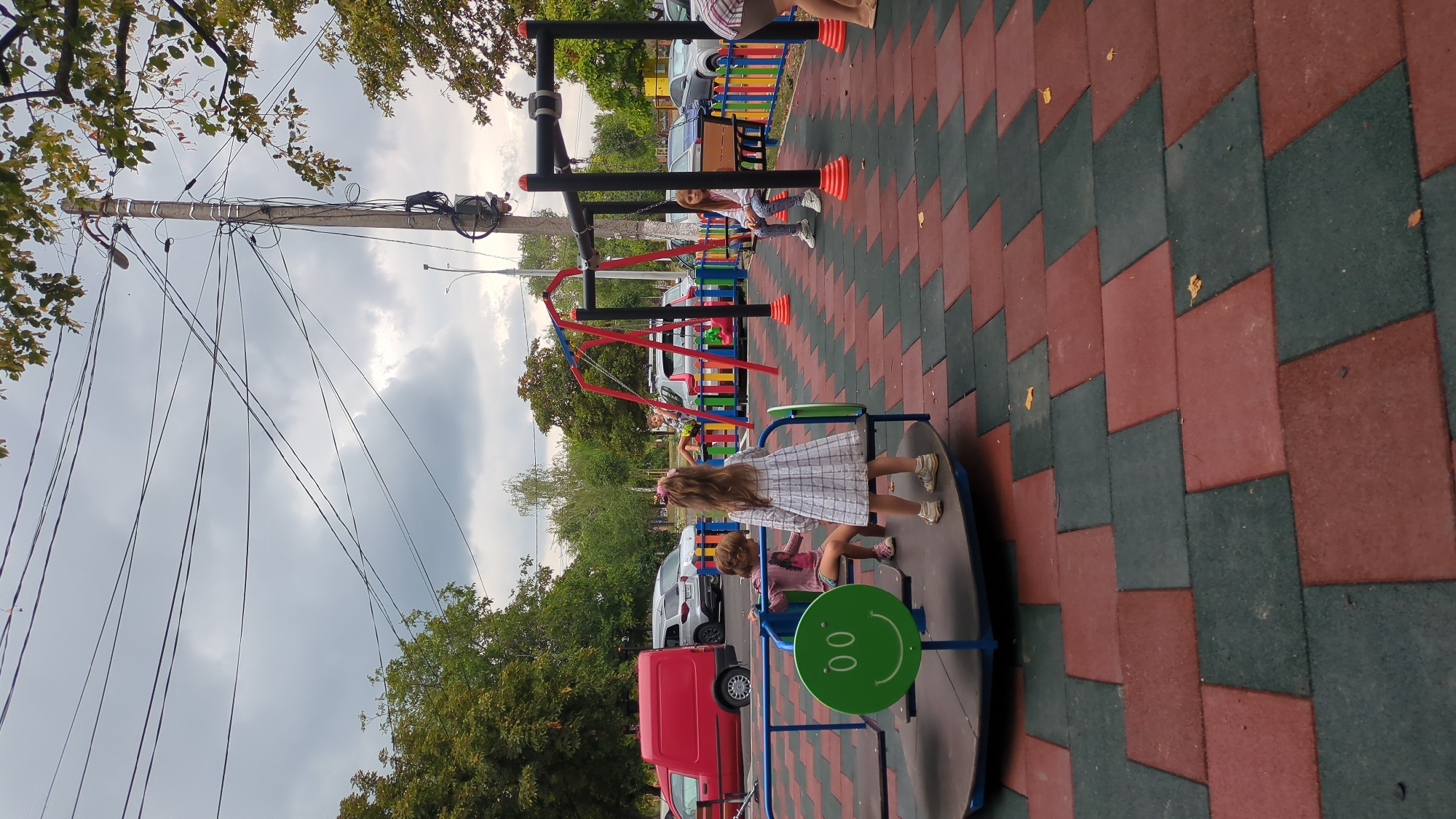 A children's playground with red and green rubberized floor tiles is depicted. Several children are present. In the foreground, a young girl in a plaid dress stands near a merry-go-round with a smiley face on its side. Another child is seated on the merry-go-round. In the background, a swing set with red supports is visible. A girl is seated on a bench under the swing set. Various cars are parked behind a colorful fence. A utility pole with numerous wires is prominent in the background, along with trees and a cloudy sky. The scene appears to be in the city of Chișinău, Moldova.