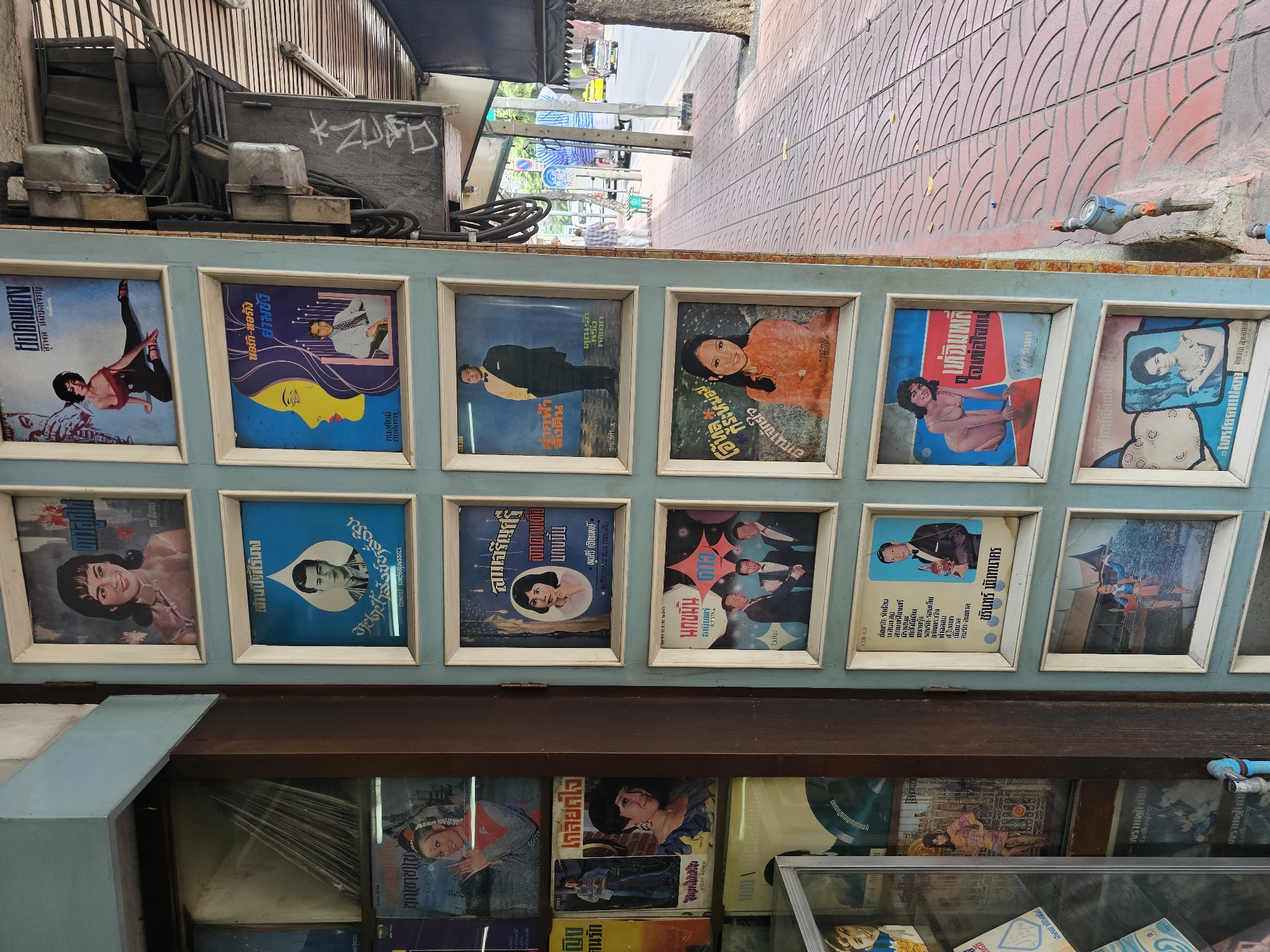 The image shows a storefront in Bangkok, Thailand, with several framed record covers displayed on a light blue wall. The record covers feature vintage images of singers and musicians, predominantly women, in colorful attire and poses. The covers have Thai script on them, suggesting they are related to Thai music from a past era. Behind the record covers, to the left, is a glass display case containing more records and possibly other merchandise. The storefront is located on a sidewalk paved with red, patterned tiles. To the right of the storefront, there is a street with vehicles, including a tuk-tuk, indicating urban activity. Electrical conduits and junction boxes are visible on the wall of the building, a common sight in many Thai cities. The overall impression is that of a vintage music shop or a stall selling old records.