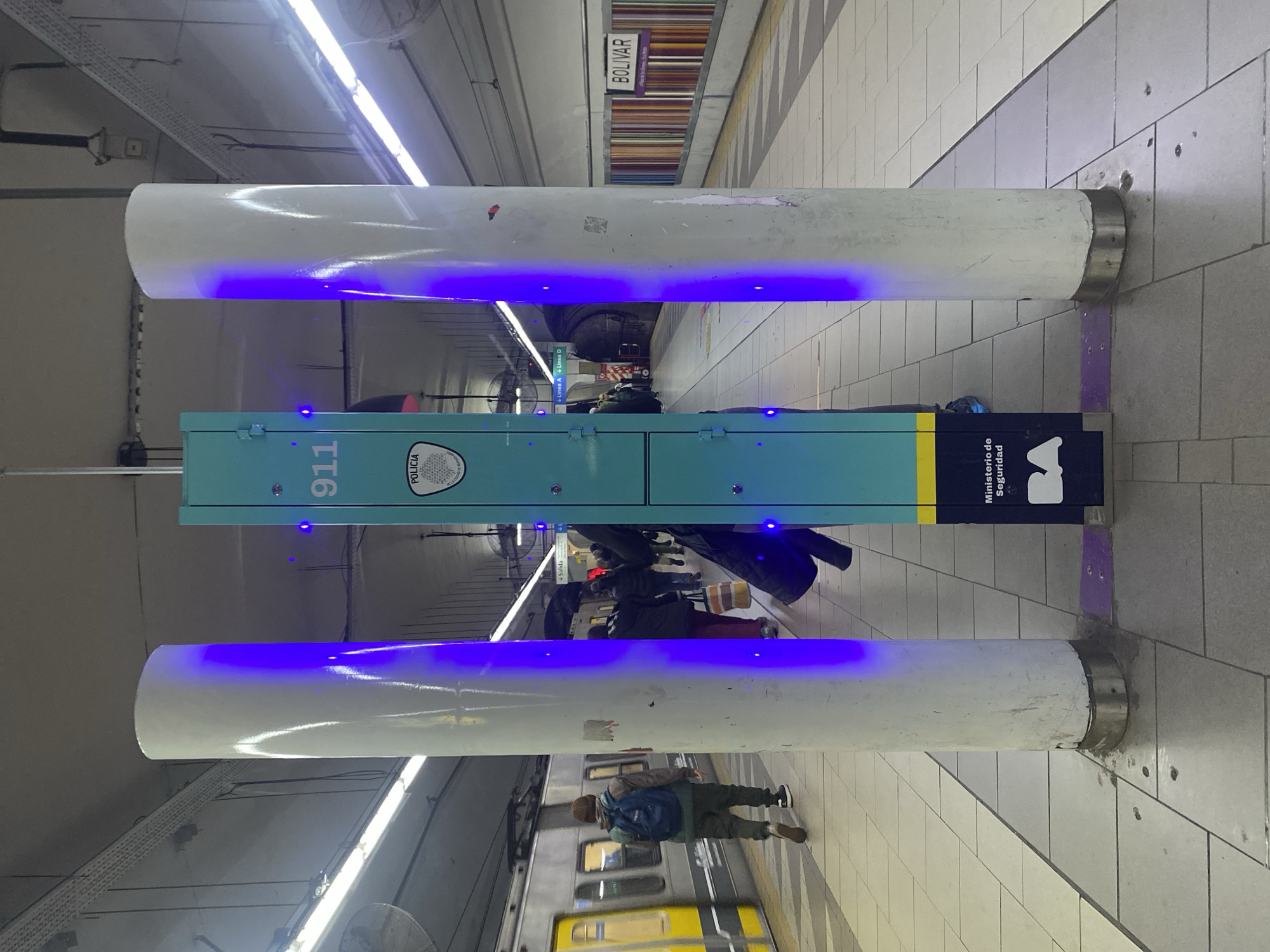 The image shows an indoor scene, most likely a subway station platform in the city of Buenos Aires, Argentina. A blue-green pillar with the number 911 and the Argentine Police logo stands between two white cylindrical structures emitting blue light. People are standing around the pillar, some waiting, and a train appears to be approaching or departing. A sign for the "BOLIVAR" station is visible overhead. The floor is tiled. The scene is well-lit with fluorescent lighting.