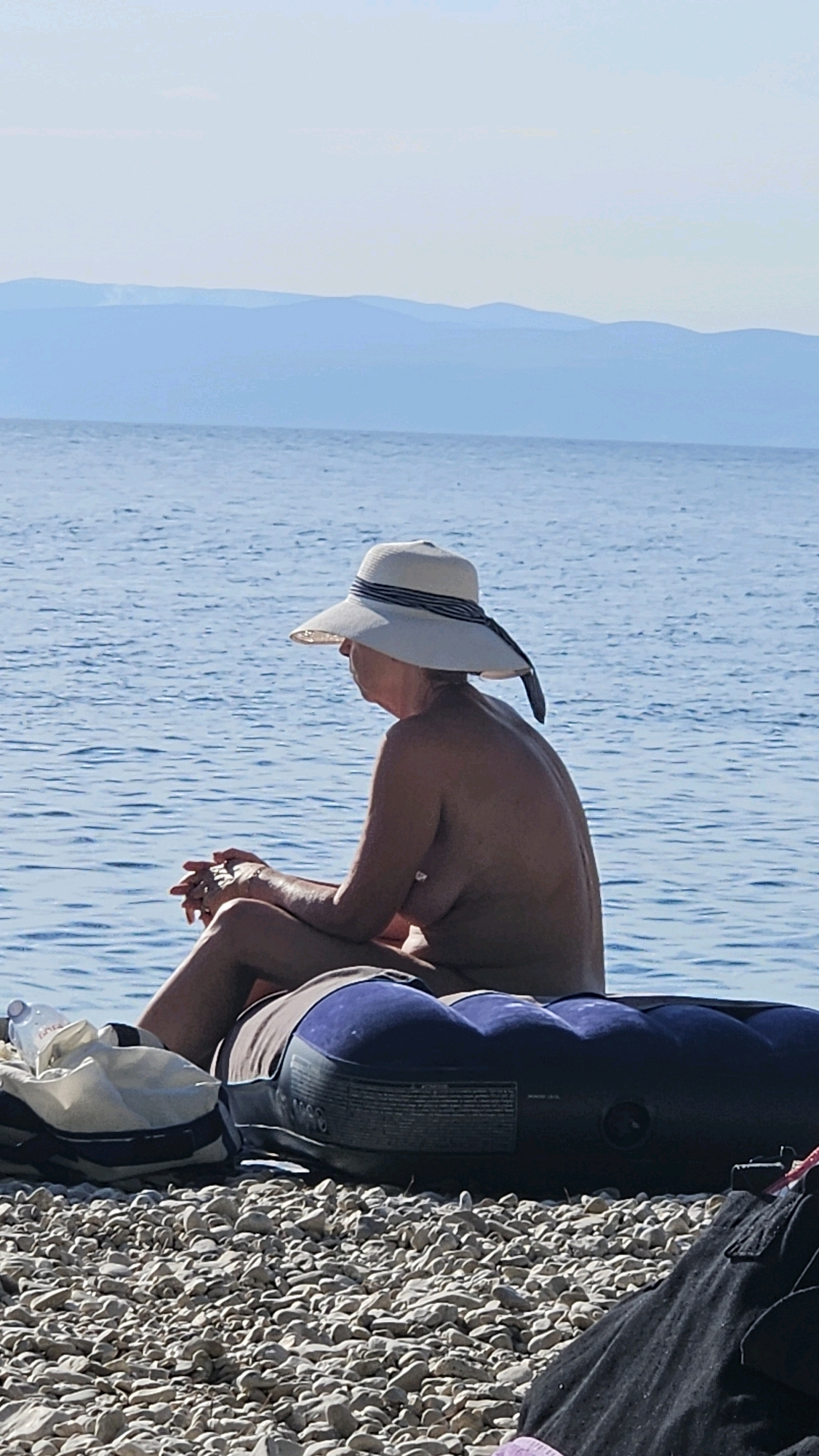 A medium shot captures a woman from the side, seated on a dark blue inflatable on a pebble beach. She wears a wide-brimmed straw hat with a dark blue and white striped ribbon. Her hands are clasped in her lap, and a bracelet is visible on her wrist. The sea, a deep blue, stretches out behind her, with faint, layered blue mountains visible on the horizon under a pale sky. In the foreground, on the left, a white and blue striped bag with a plastic bottle on top rests on the pebbles. To the right, dark fabric, possibly clothing, is visible on the ground. The lighting suggests it is a sunny day.