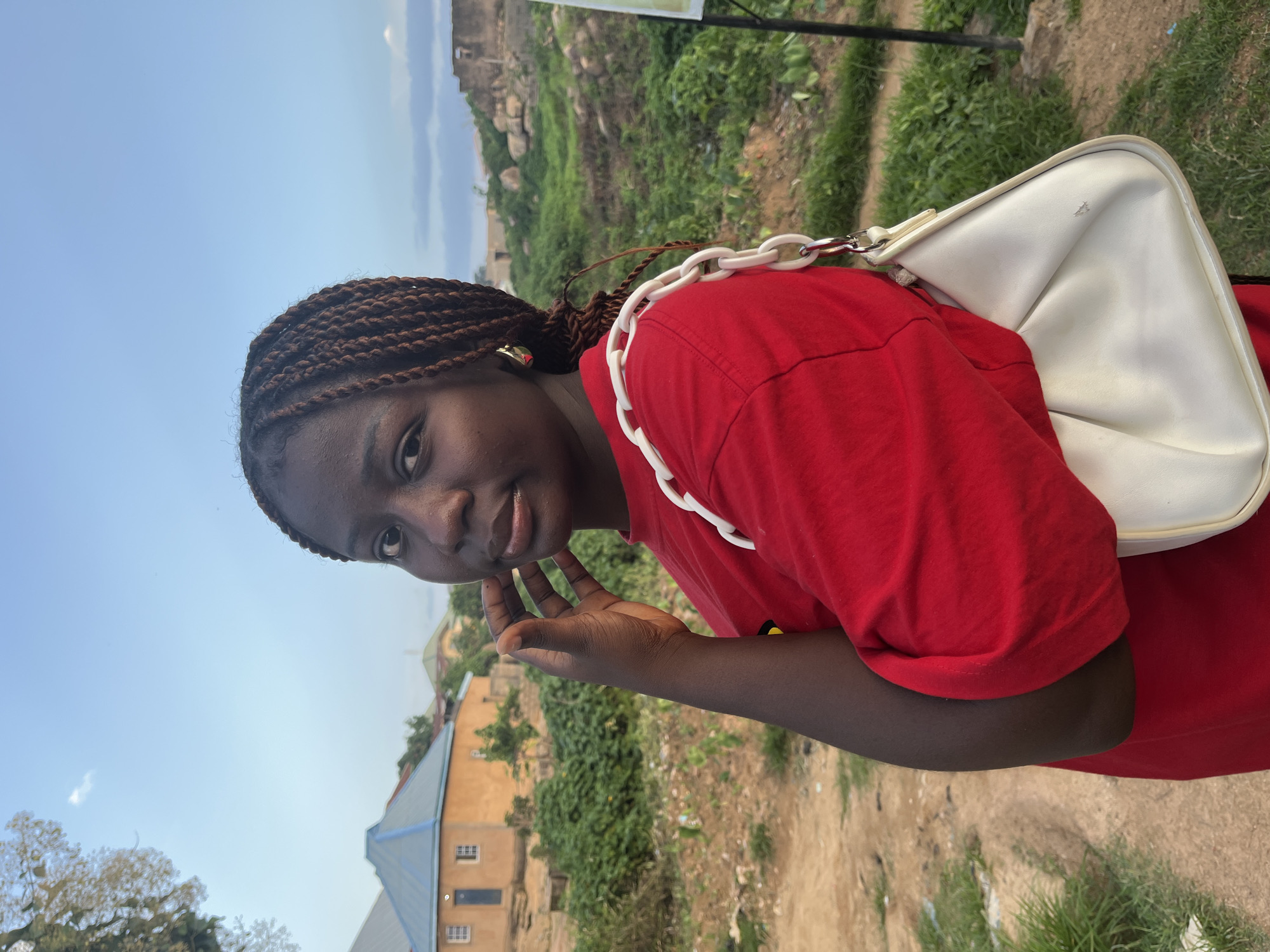 A medium shot captures a young woman with dark skin and braided hair from the chest up, facing left with her head turned towards the camera. She wears a red t-shirt and a white handbag with a thick white chain strap is slung over her left shoulder. Her right hand rests on her chin, and she looks directly at the viewer. The background features a dusty, uneven ground with scattered green vegetation and a few buildings, including a house with an orange exterior and a blue roof, indicative of a rural or semi-urban setting. The sky above is a clear blue with a few wispy clouds. The overall lighting suggests it is late afternoon or early evening.