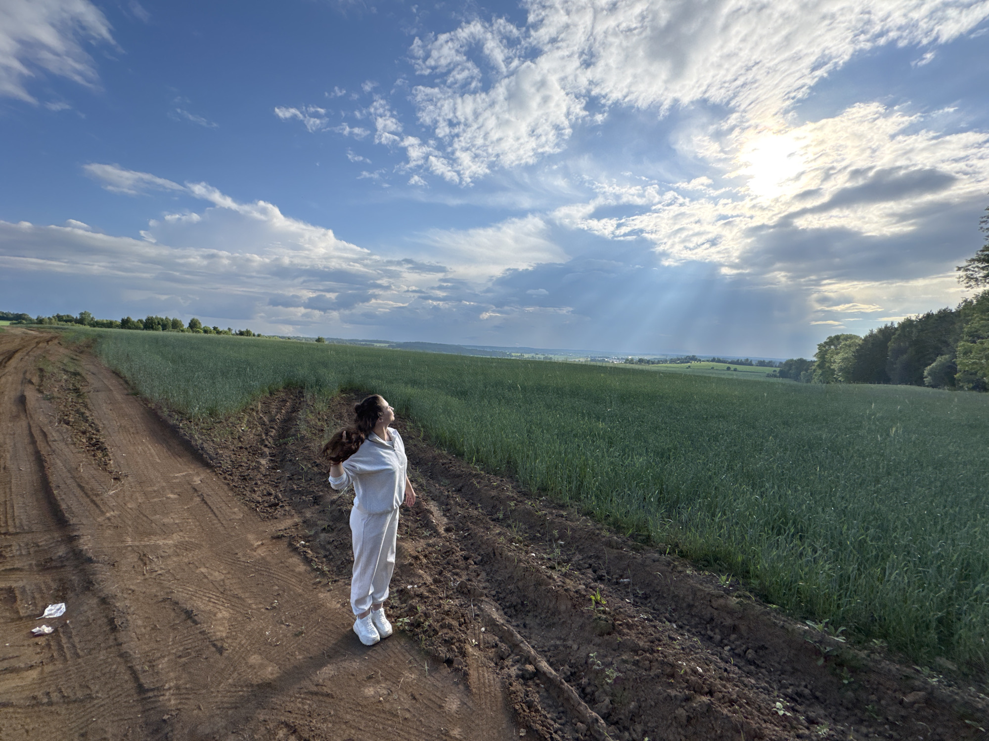 A young woman in a light-colored tracksuit stands on a dirt road in a field, looking up at the sky. The road is to her left, and a green field of crops stretches out to her right. The sky is blue with dramatic white clouds, and the sun is partially obscured by clouds, casting rays of light onto the landscape. In the distance, trees line the horizon. The overall scene suggests a rural or agricultural setting, possibly in or near Lukintsy, Russia.