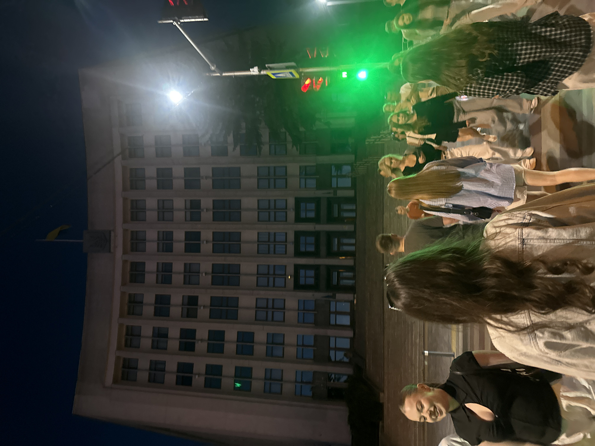 In Ternopil, Ukraine, at night, a group of young people are walking on a street in front of a large, brutalist-style building with many windows. A yellow flag flies from the top of the building. A traffic light is visible, displaying a green signal. The people are dressed in casual attire. One woman in the foreground on the left is smiling. The scene appears to be outdoors on a city street.