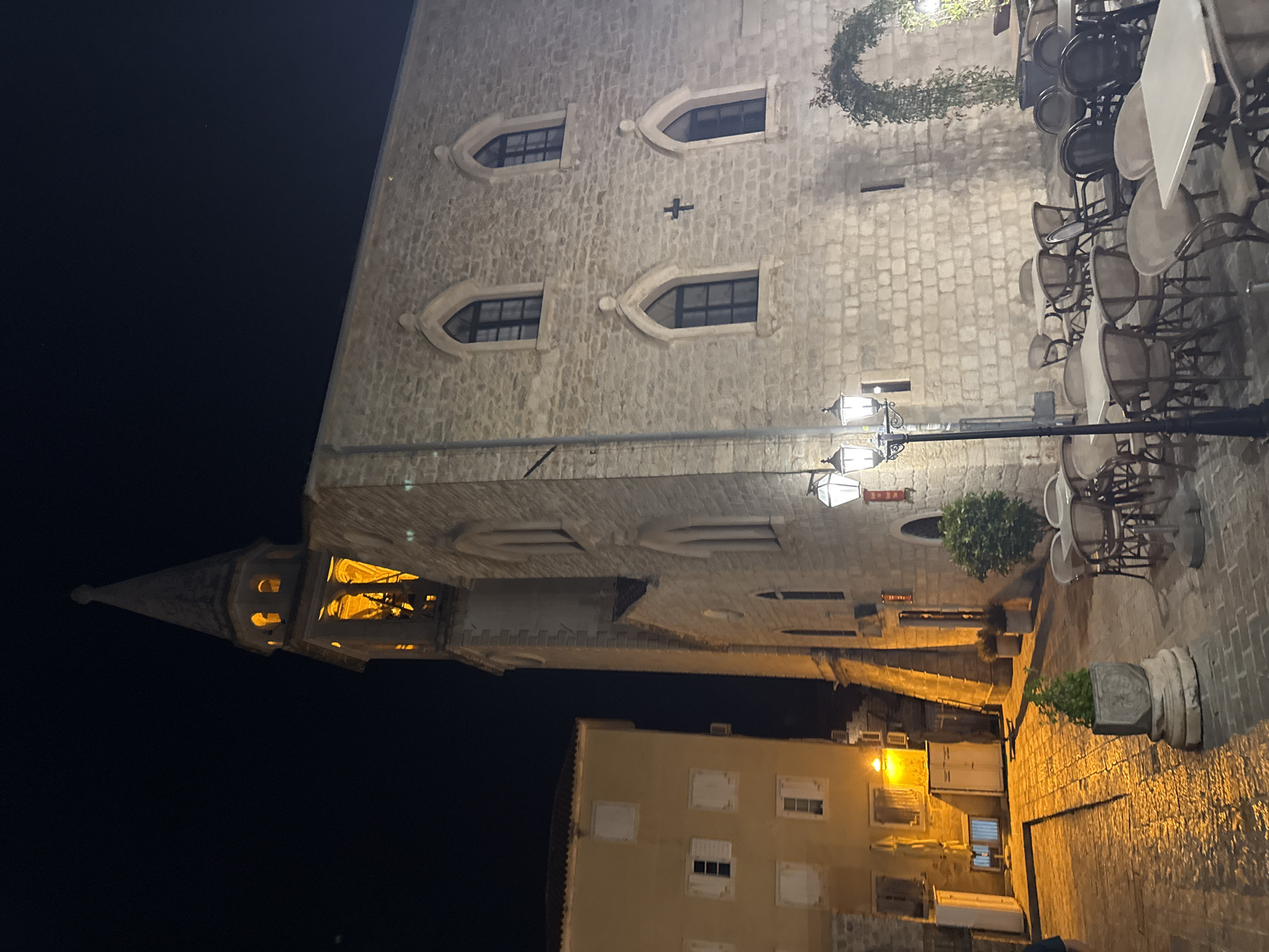 The image shows a nighttime view in Budva, Montenegro, featuring the exterior of a historical building with stone walls and arched windows. A church steeple is visible at the left. Street lighting illuminates the scene, and outdoor seating is arranged on the right side of the image, suggesting a cafe or restaurant. The sky is dark.