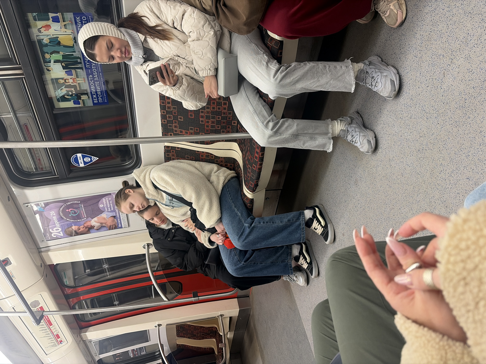 A photo taken inside a Saint Petersburg, Russia, metro car shows several people sitting. One woman wears a white quilted jacket and hat, light gray jeans, and white sneakers and looks at her phone. Two other women are asleep next to each other, and both wear jackets. A sign for a clinic advertises reproductive health and is displayed on the wall of the car. A second sign promotes courtesy towards children and passengers with disabilities.