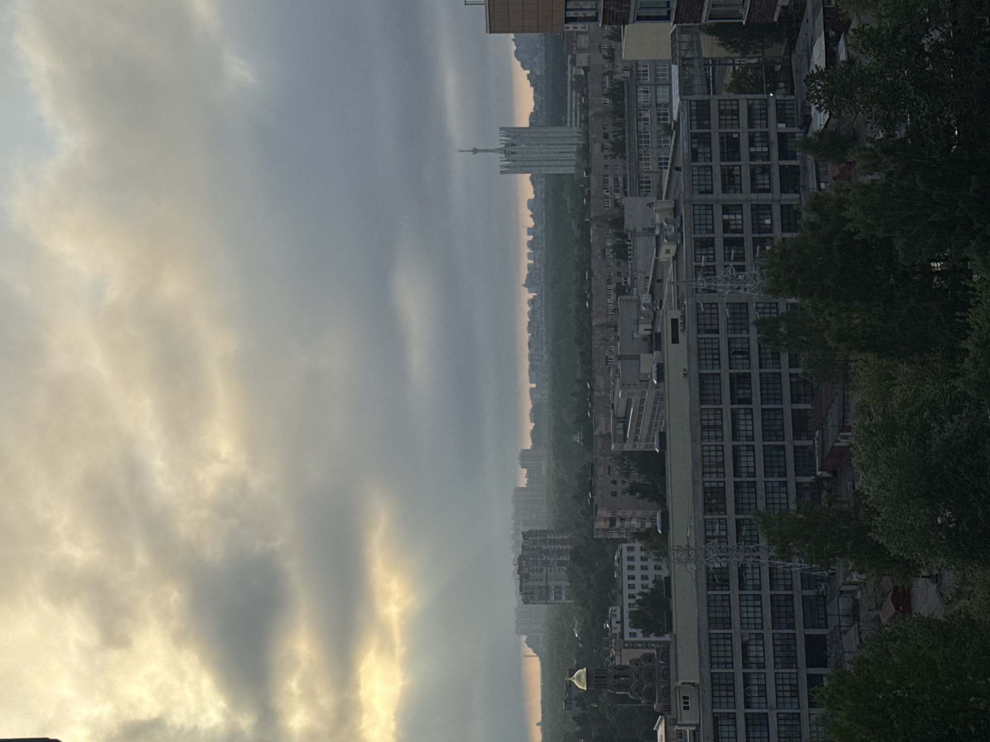 The image shows a hazy cityscape, likely taken from a high vantage point in Sinyavino, Russia. The sky is filled with overcast clouds, some tinged with a golden hue, possibly from the setting or rising sun. Below the clouds is a dense urban landscape with a mix of buildings, ranging from low-rise structures to taller apartment blocks. A large, prominent tower with a spire can be seen in the distance. Closer to the viewer, a series of grid-like apartment buildings dominate the view, with trees lining the streets and some encroaching on the buildings. There are no visible people or discernible actions. The environment appears damp or misty, contributing to the overall atmospheric haze.