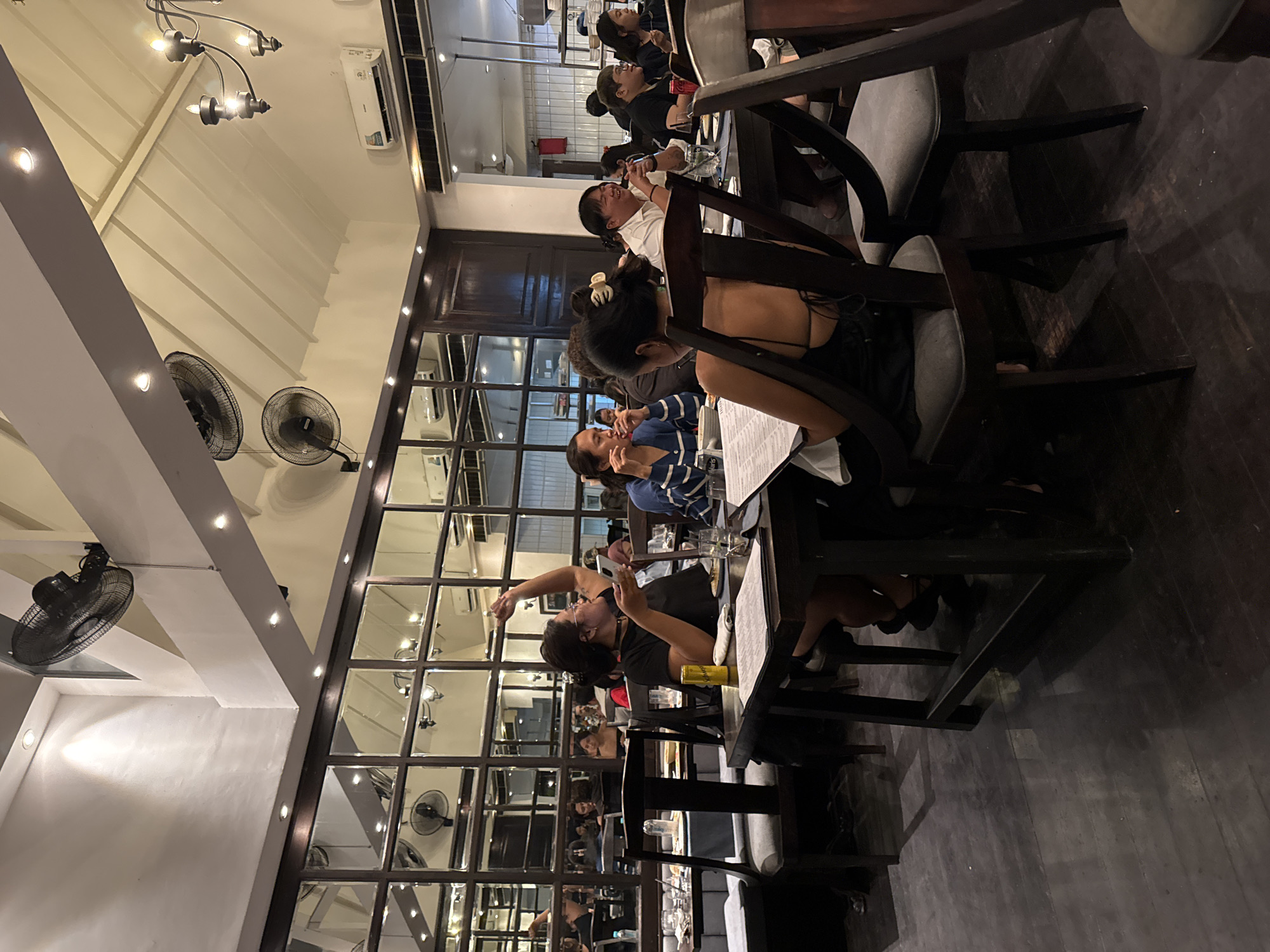 The image captures an indoor scene at a restaurant in North Kuta, Indonesia, likely at night, given the artificial lighting. Several groups of people are seated at dark wooden tables, engaged in dining and conversation. A prominent feature of the restaurant's interior is a large wall of windows with dark frames, which appear to be mirrored on the interior, reflecting the dining area and ceiling. The ceiling is sloped and has exposed beams, with industrial-style ceiling fans mounted on the upper walls. Overhead lighting fixtures, including a chandelier, illuminate the space.
In the foreground, a woman in a black top with spaghetti straps is seated at a table, holding a mobile phone and gesturing with her right arm. A yellow can, possibly of a beverage, sits on the table in front of her. Other diners are visible at adjacent tables, some interacting with each other, while others appear to be focusing on their food or phones. The flooring is dark wood, with reflections from the lighting. The overall ambiance suggests a lively dining establishment.