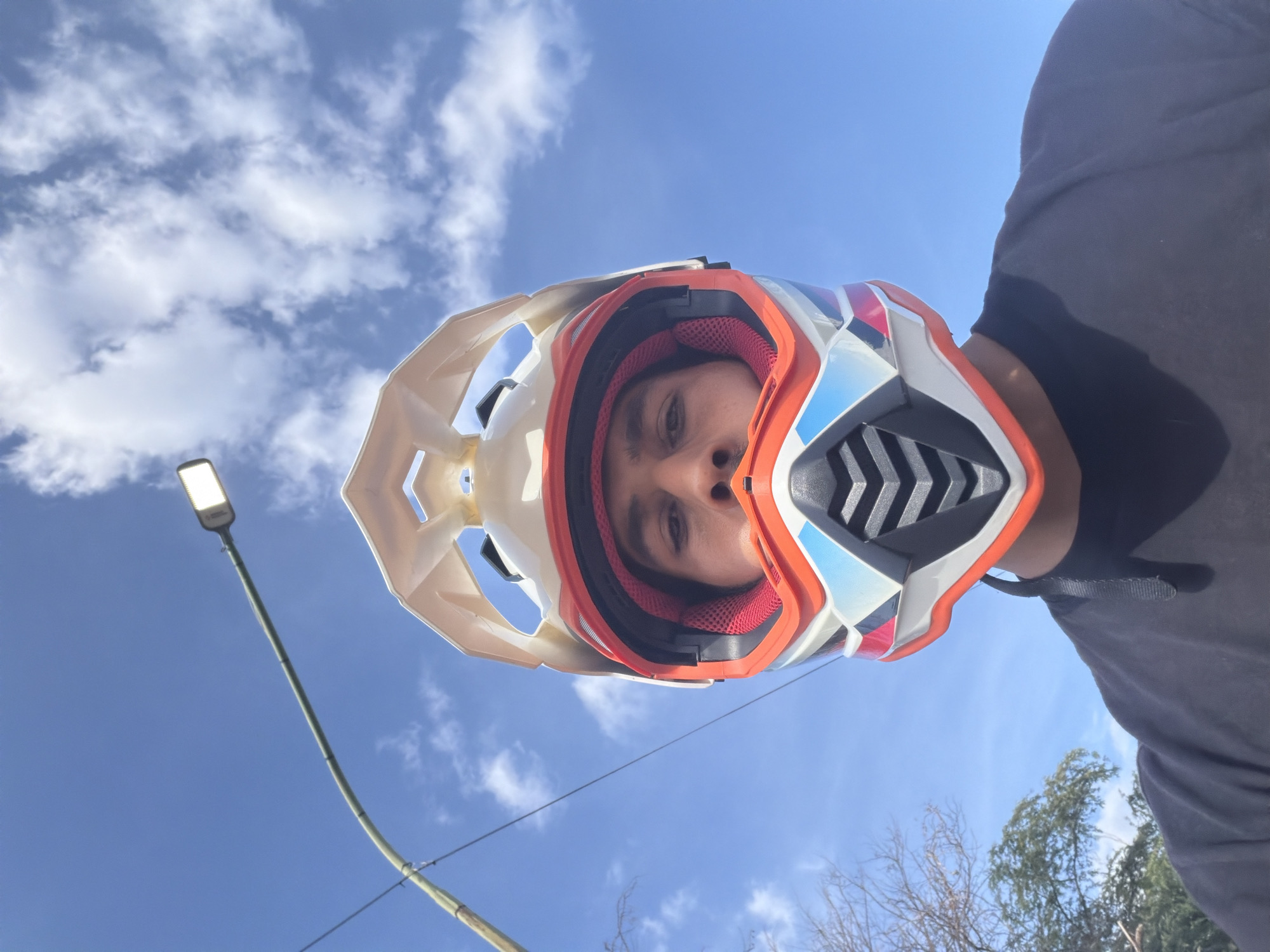 A low-angle, close-up, outdoor, eye-level, waist-up, portrait-oriented, eye-level, selfie-style shot features a person wearing a white, orange, red, and blue motocross helmet, facing the camera. The helmet's visor is visible, and the person's eyes and nose are peeking out from the opening. They are wearing a black t-shirt. The background consists of a clear blue sky with scattered white clouds. A street light on a curved pole extends from the left side of the frame towards the upper right. Trees and foliage are visible in the lower left corner. The scene appears to be taken during the daytime, with bright sunlight illuminating the subject. The location is identified as Turmero, Venezuela.