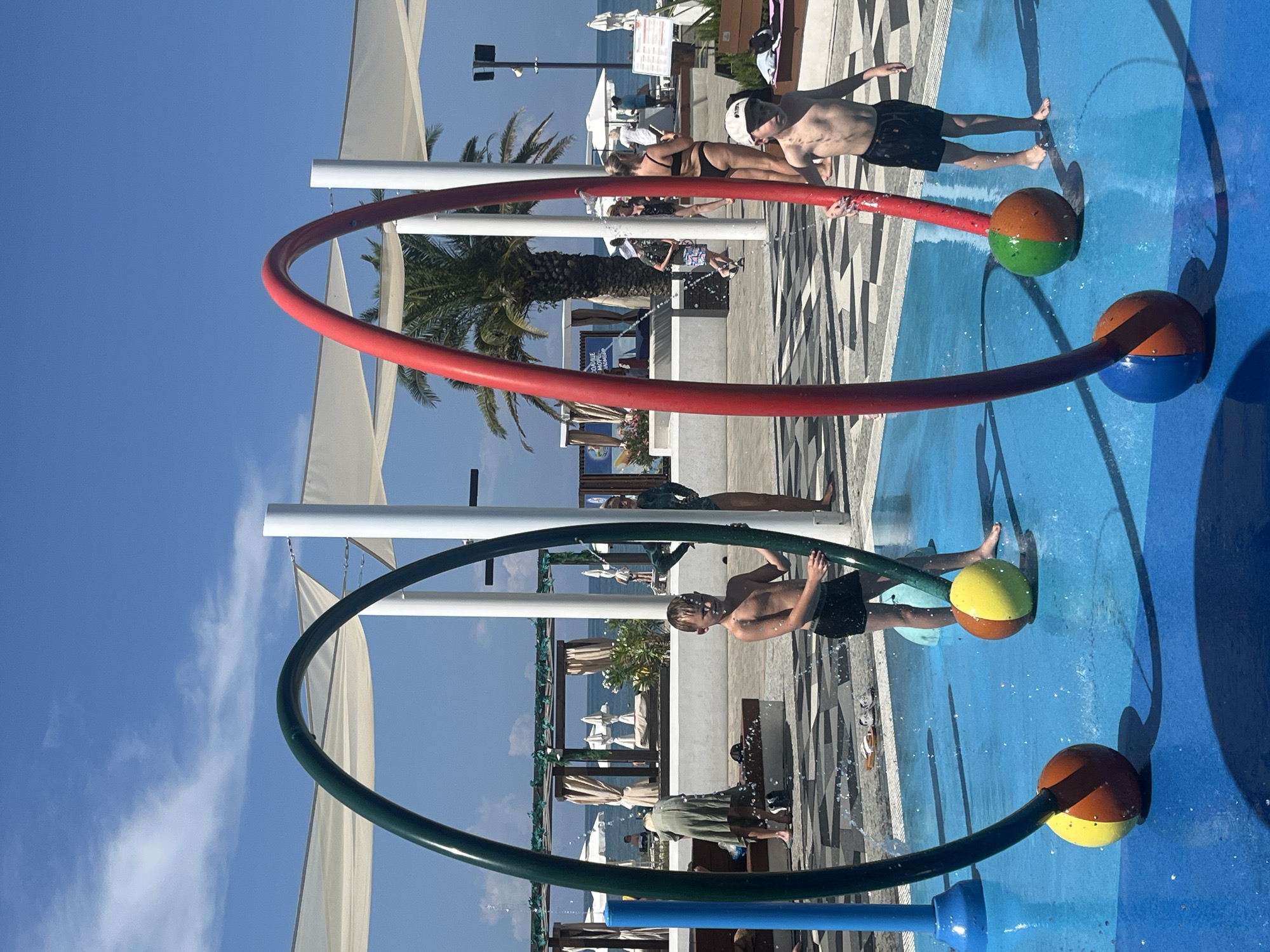 The image shows a shallow water play area, likely a splash pad, with several people enjoying the water. The scene is set outdoors on a sunny day, with a clear blue sky and a few white clouds. Palm trees are visible in the background, suggesting a warm climate. The water play structure consists of two large, colorful rings, one red and one green, with spray nozzles. Attached to the rings are spherical sprinklers that release water.
In the foreground, two young boys are playing in the water. One boy, wearing black swim trunks, is standing near the green ring, with water spraying around him. He appears to be laughing or smiling. The other boy, also in black swim trunks, is further back near the red ring. Another person, an adult male, is also visible in the background, seemingly interacting with the water feature. Further in the background, there are lounge chairs and umbrellas, indicating a resort or hotel setting near a body of water, possibly the sea.
The overall mood of the image is joyful and playful, capturing a moment of summer fun. The lighting suggests it is daytime, likely late morning or early afternoon. The presence of a palm tree and the blue sky are strong indicators of a warm weather location. There is no visible text that can be clearly discerned in the image. The reflection of the people and the structure in the wet surface of the splash pad adds depth to the scene.