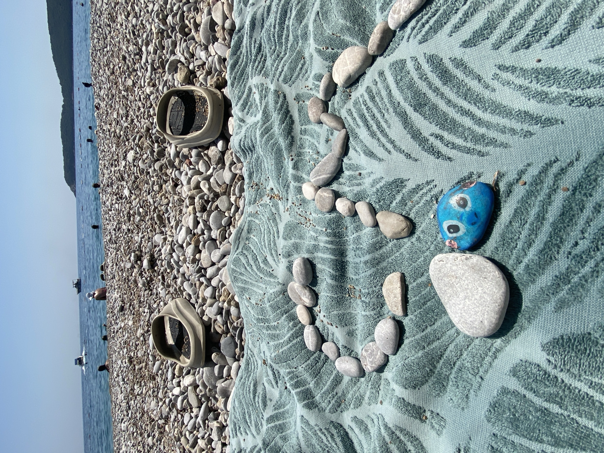 A high-angle, medium shot shows a beach scene in Prijevor, Montenegro, on a clear, sunny day. The foreground features a teal-colored towel with a palm leaf pattern. On the towel, a line of smooth, grey stones forms a curve, resembling a fish's tail or body. A small, blue-painted rock with large, cartoonish eyes and pink cheeks is placed to the left of the stone formation, appearing to be the head of the fish. To the right of the blue rock is a larger, grey, oval-shaped stone.
In the middle ground, a pebbly beach stretches towards the sea. Two tan-colored flip-flops, upside down and filled with pebbles, are placed on the beach, one on each side of the frame. The sea is calm and blue, with several boats visible in the distance. A few people are in the water or on the beach, some appearing to swim. The background consists of a treed hillside.