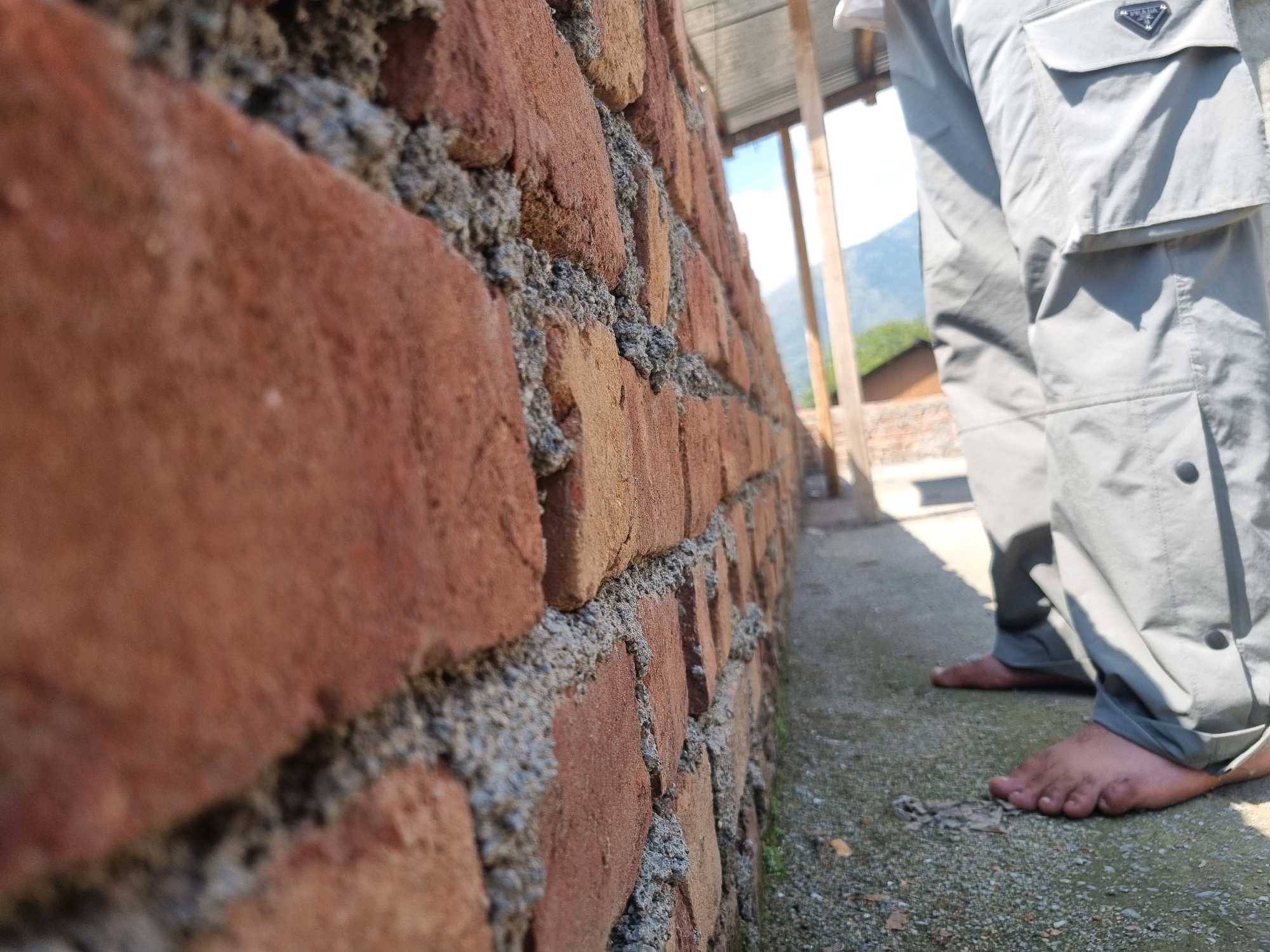 In Manigam, India, a close-up, low-angle shot focuses on a rustic brick wall with visible mortar, suggesting it's outdoors. To the right, a person's lower body is visible, clad in light gray cargo pants with black snap buttons. The person is barefoot, with their left foot closest to the camera, resting on a gravelly ground. Sunlight casts shadows from overhead structures, and the background hints at a mountainous landscape with trees and buildings. The scene appears to be in a casual, possibly rural or semi-urban outdoor setting within Manigam.
