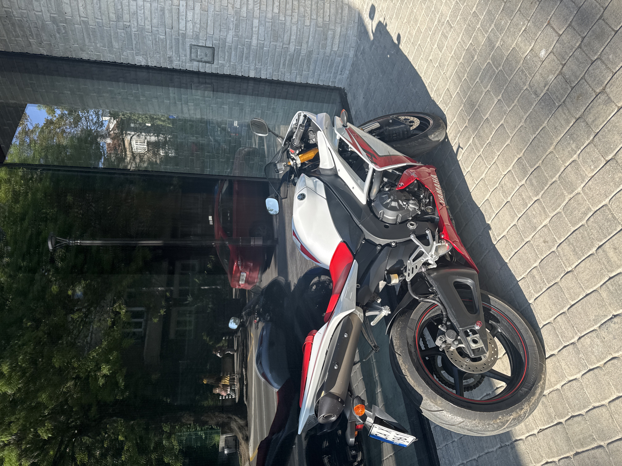 A red, white, and black Yamaha motorcycle is parked on a cobblestone sidewalk beside a modern building in Sofia, Bulgaria. The building has a gray brick wall and large glass windows that reflect the surrounding urban environment, including a red car, trees, and other buildings. The motorcycle is positioned at an angle to the building. A shadow is cast across the sidewalk. The motorcycle's license plate is visible.