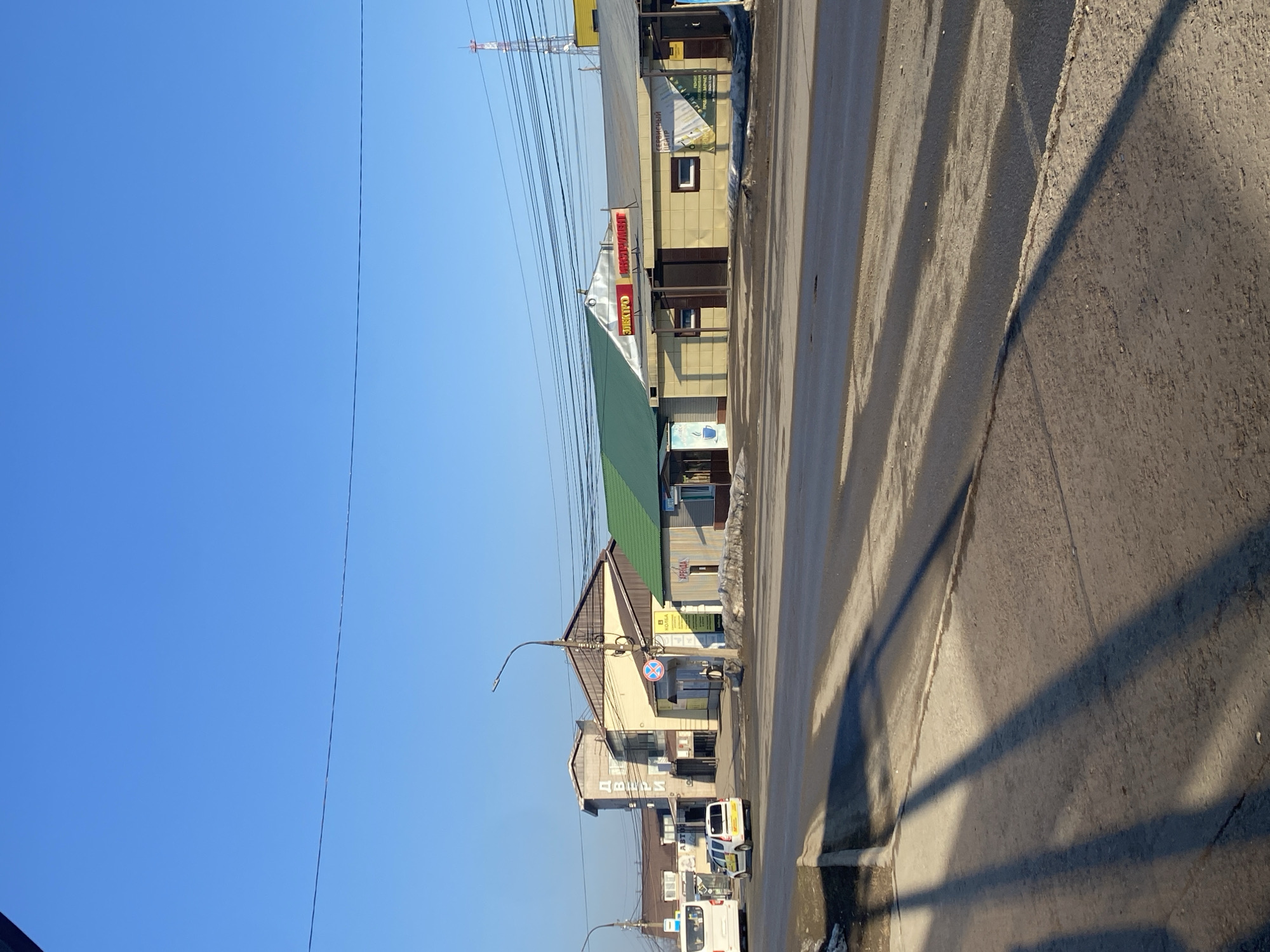 The image captures a street scene in an urban area. The sky is clear and blue. Buildings line the street, including one with a green roof. Power lines are visible overhead. A road or sidewalk runs along the right edge of the image, with shadows cast across it. Vehicles are present on the street. The image appears to be taken from a standing position.