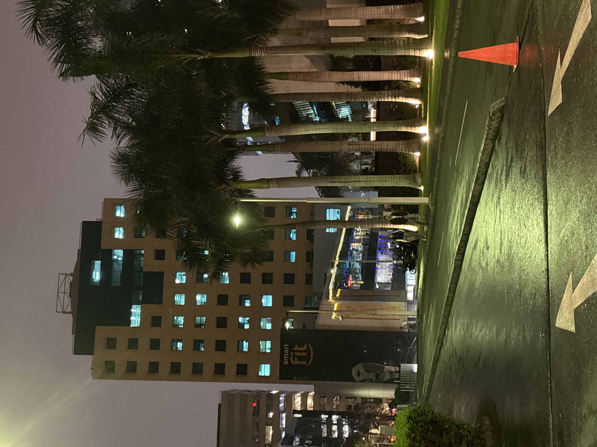 At night, in San Isidro, Peru, a multi-story building with many illuminated windows stands behind a row of tall palm trees. In front of the building, there is a large black advertisement for "Smart Fit." To the left of the advertisement, a sculpture is visible. A street runs diagonally across the foreground, wet and reflecting the lights. On the right side of the street, an orange traffic cone is positioned. Two individuals are walking on the sidewalk near the Smart Fit advertisement, and a parked motorcycle is visible behind them. The sky is overcast and dark.