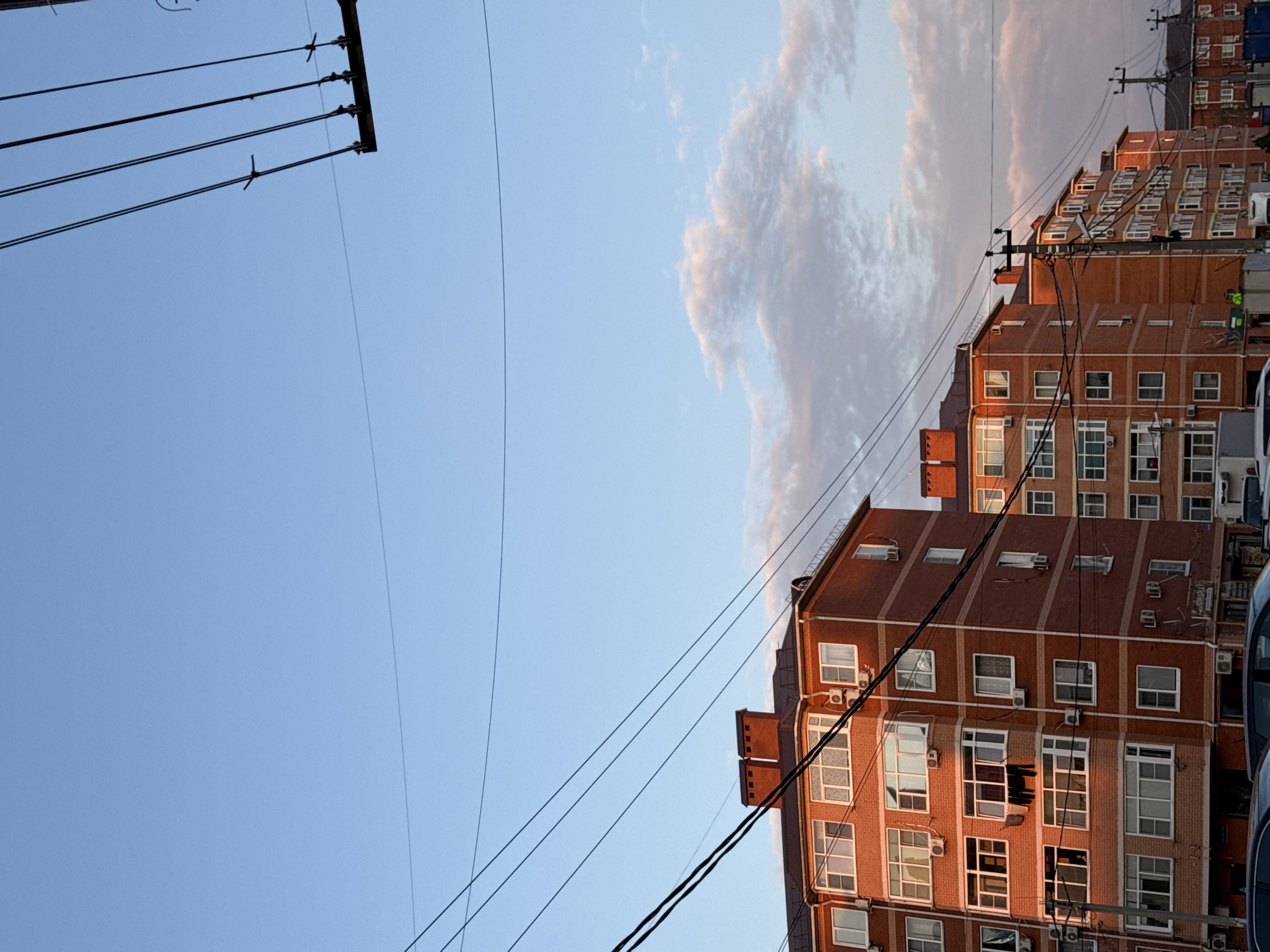 The image captures a view in an urban setting. The sky, occupying a significant portion of the frame, is a gradient of blue with a smattering of clouds tinged with light pink. A set of power lines runs across the sky, anchored by a dark-colored utility pole in the upper left corner.
On the right side of the frame, a brick building dominates the view. The building is multi-storied with numerous windows and air conditioning units. The facade is bathed in the warm light of the setting or rising sun, casting long shadows. The architecture is modern, but the building shows signs of age and wear. The image is rotated sideways, giving an unusual perspective of the building rising almost vertically.