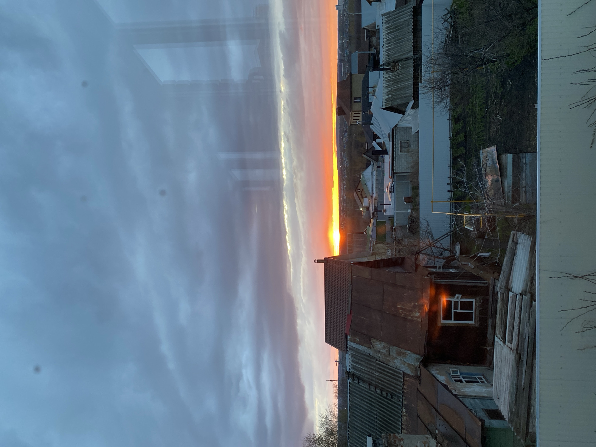 The image, displayed vertically but depicting a horizontal landscape, shows a sunrise or sunset over a residential area in what is likely Ulyanovsk, Russia, viewed from an interior window. The sky dominates the upper half, featuring long, horizontal streaks of clouds. A vibrant band of intense orange and yellow light, indicating the sun's position, stretches across the horizon, transitioning into pale orange, grey, and darker blue-grey clouds higher in the sky.
Below the horizon, a collection of residential rooftops and structures fills the foreground and midground. Many roofs are made of corrugated metal, some exhibiting rust or wear, alongside tiled or flat roofs. The buildings vary in design, with a utilitarian aesthetic. Bare tree branches are interspersed among the structures, suggesting a colder season like late autumn or early spring. A prominent window on a dark-colored house in the foreground reflects the bright light of the sky. A yellowish utility pipe is also visible.
The photograph was taken from indoors, indicated by reflections of what appear to be window frames and faint interior elements on the left side. Several dark, circular smudges are visible on the windowpane. No people or visible text are present. The scene conveys a sense of quiet and tranquility, capturing a moment of natural beauty within an urban setting.
