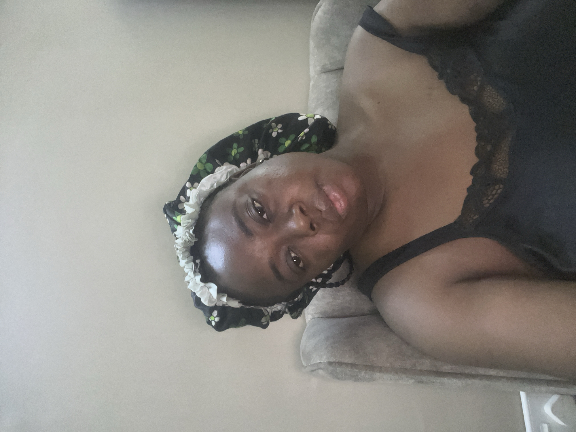 A horizontally oriented, medium close-up photograph features a person, likely a woman, with dark skin, lying down and looking directly into the camera. She is wearing a black top with delicate lace trim along the neckline and shoulder straps. Her hair is covered by a black bonnet or sleep cap, which is patterned with small green leaves and white flowers, and has a white, ruffled elastic edge. Her lips are slightly parted, and her expression is neutral and relaxed.
The background primarily consists of a plain, light grey wall. The person is resting on a grey upholstered surface, possibly a couch or bed, which is visible beneath and behind her. The lighting is soft and appears to be indoors. There are no other discernible objects or text in the image. The scene suggests a casual, indoor setting, possibly a home in Abuja, Nigeria.